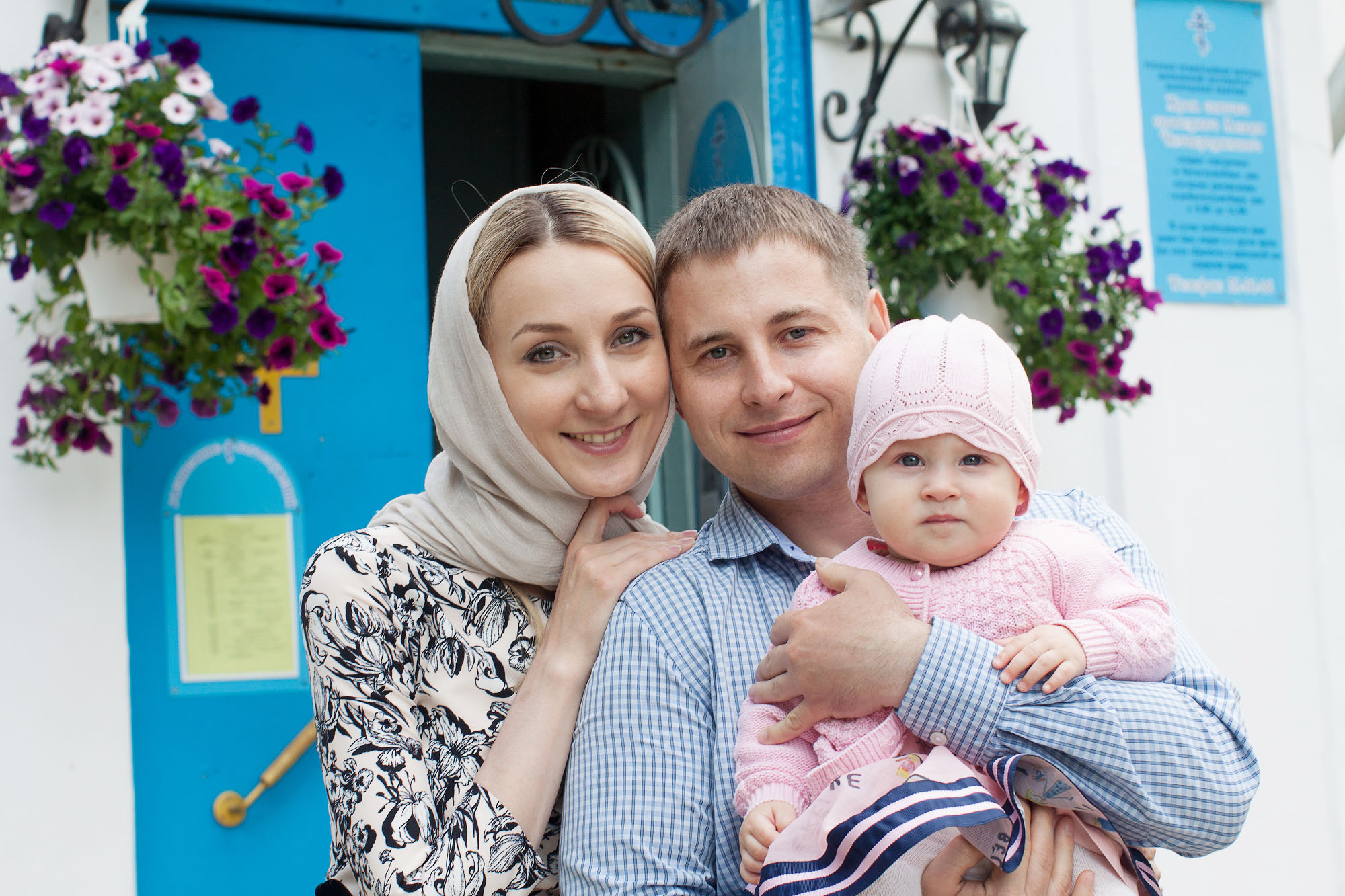 Женщина и мужчина с девочкой на руках стоят на фоне церковных дверей синего цвета.