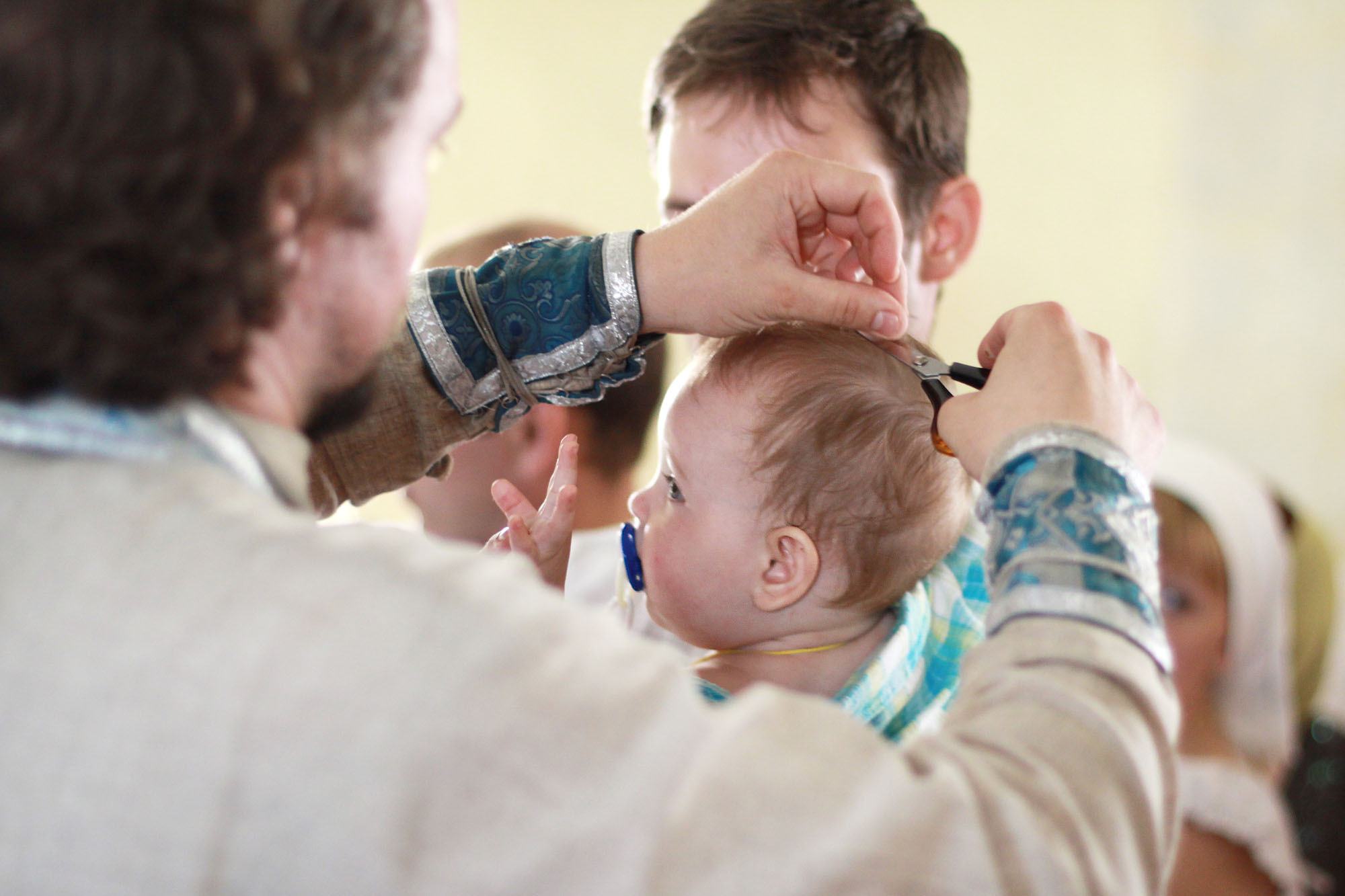 Священник в светлой рясе состригает волосы у ребенка.