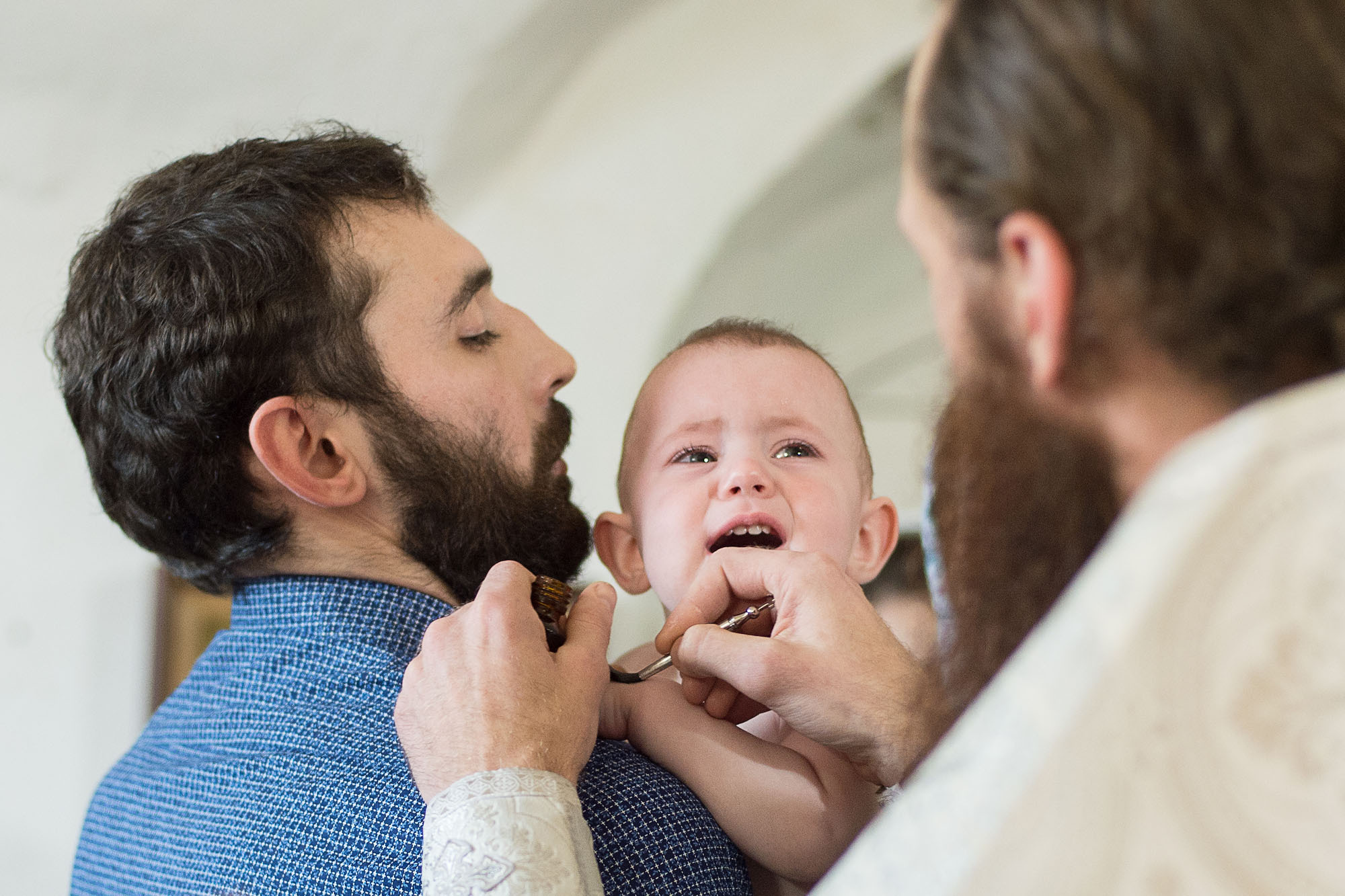 Священник делает обряд помазания малышу, который сидит у мужчины с бородой и синей рубашке.