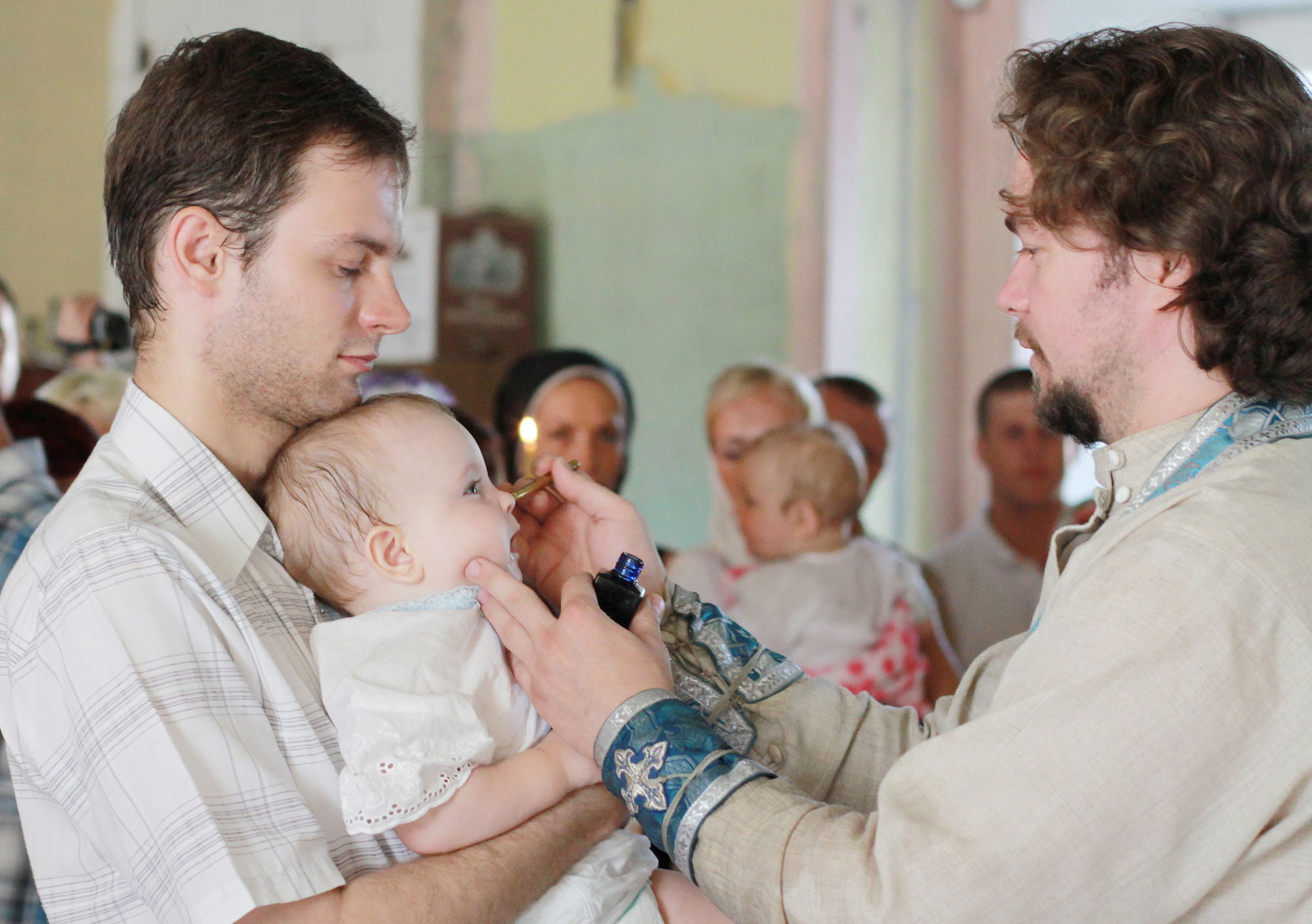 Священник в светлой рясе осуществляет обряд помазания ребенку на лице.