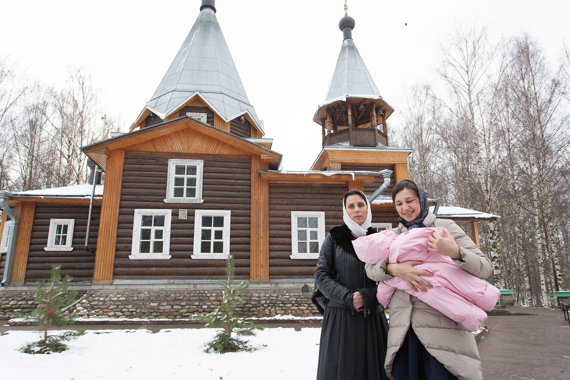 Две женщины с ребенком на руках стоят зимой на фоне деревянной церкви.