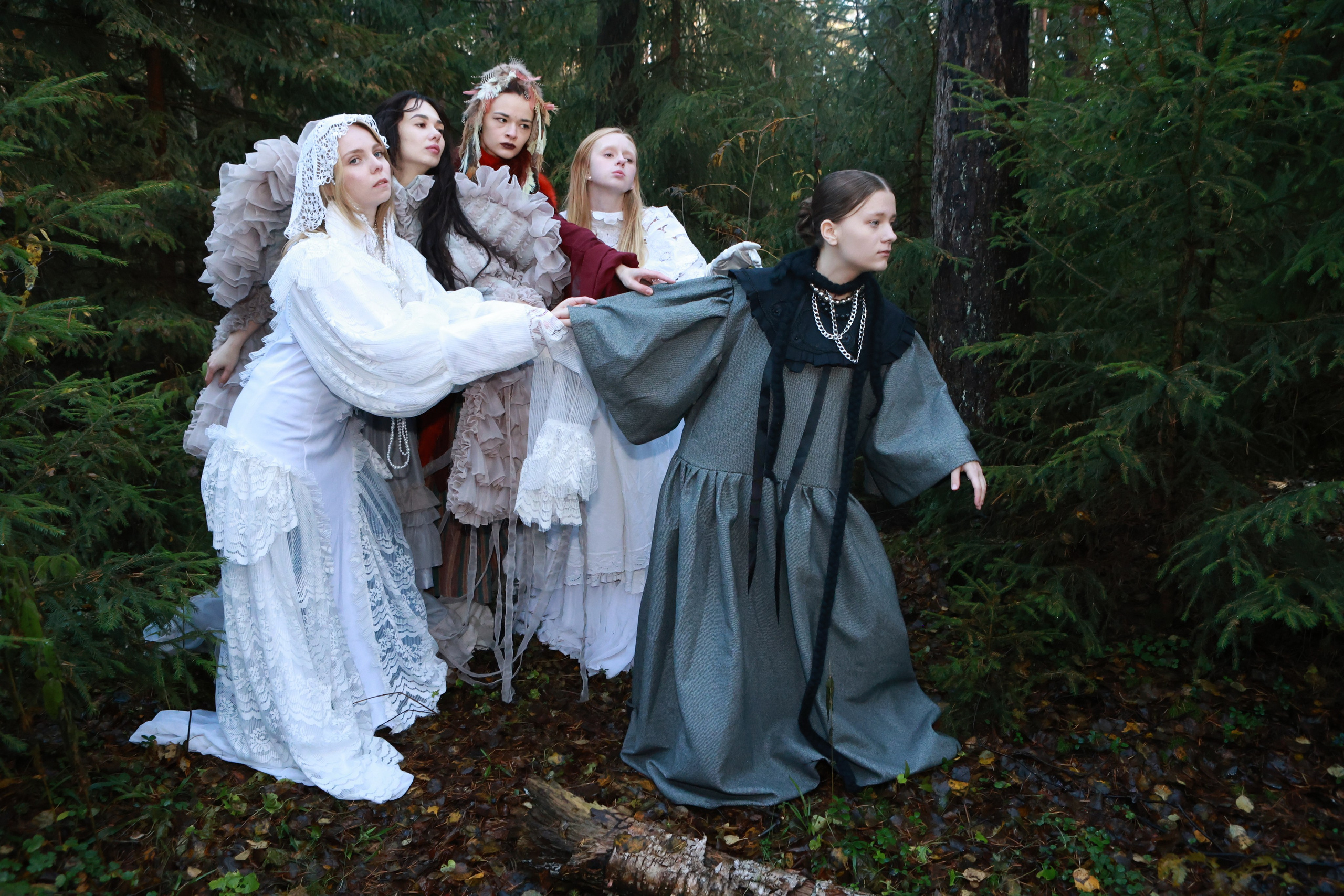 Группа из пяти моделей в винтажных платьях с кружевом и рюшами позирует в хвойном лесу. Серое платье с жемчужной цепочкой, белоснежные наряды, динамичные жесты. Осеннее настроение, ели и опавшие листья на фоне. Арт-фотосессия.