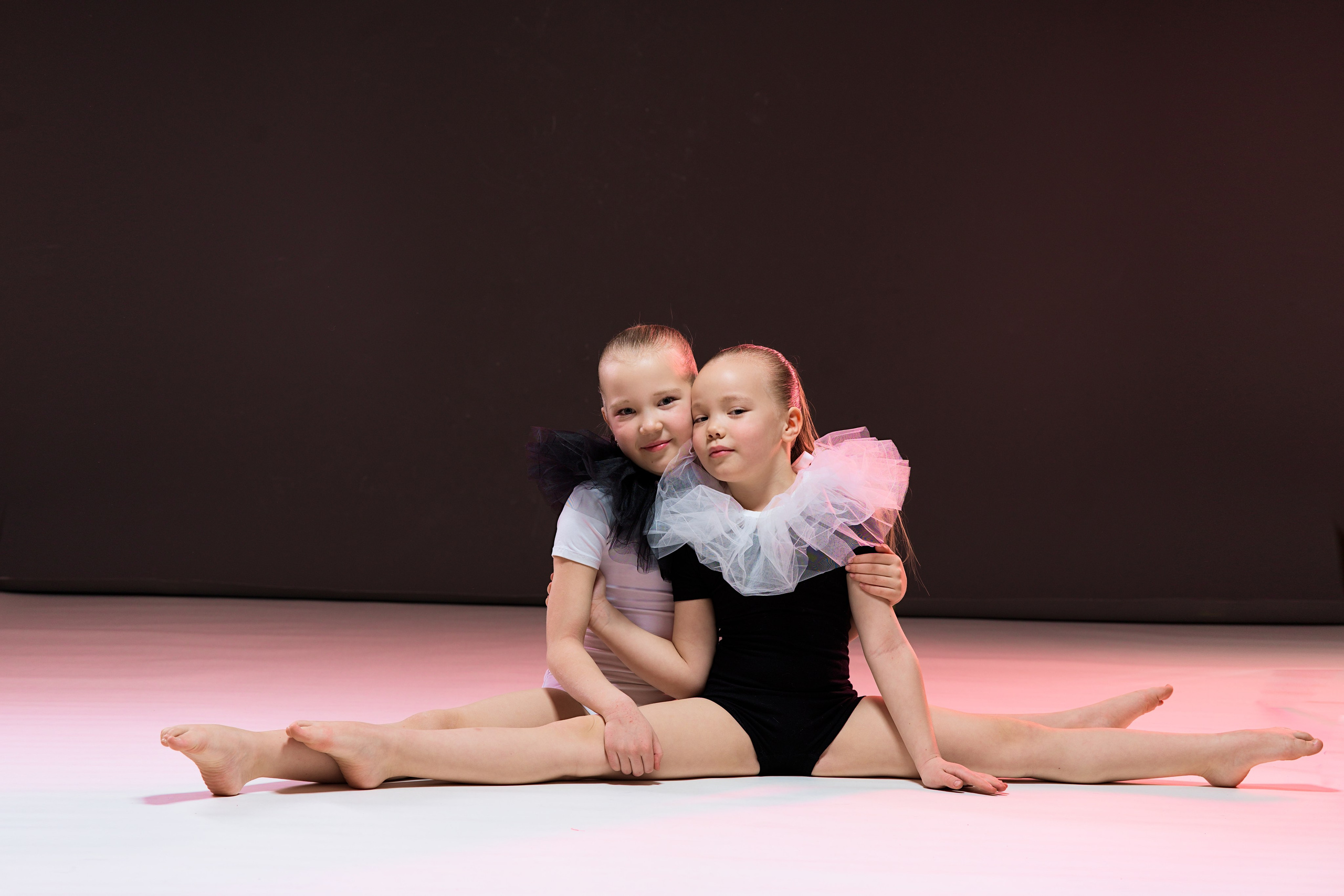 Искренние объятия двух юных гимнасток, сидящих в шпагате на полу студии. Художественная фотография, передающая атмосферу дружбы в спорте.