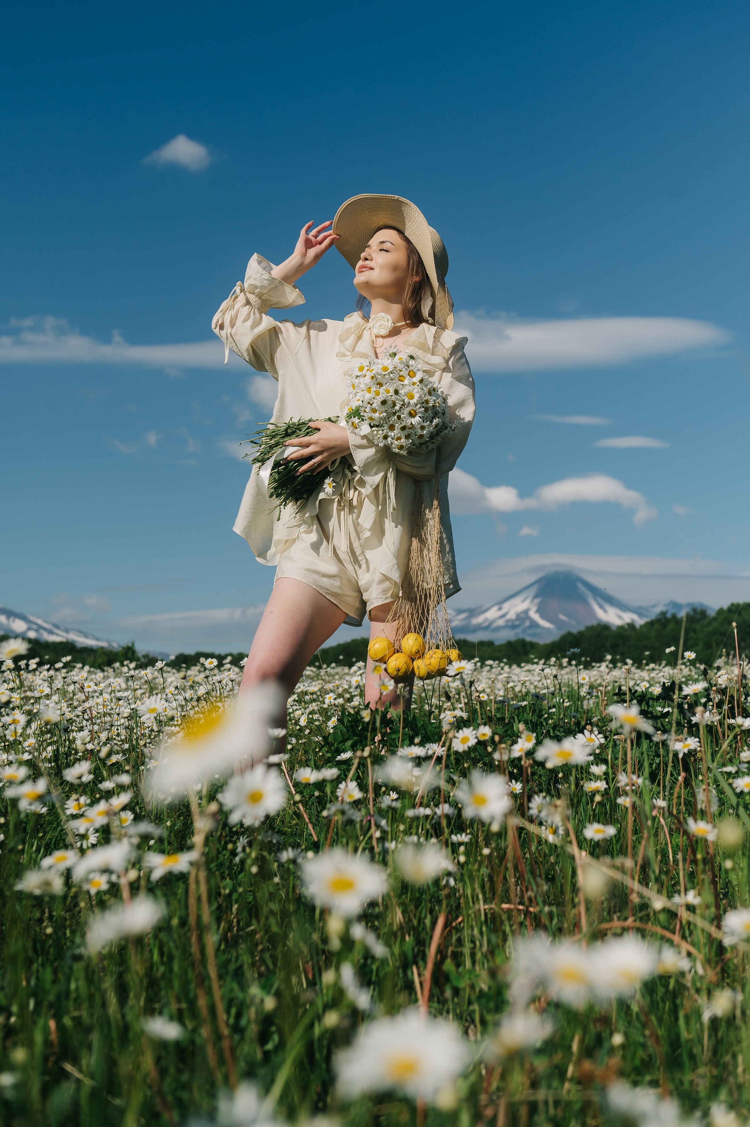 Девушка в светлой одежде и шляпе стоит в ромашковом поле, держит букет. На фоне — горы, небо ясное. Атмосфера умиротворённая.