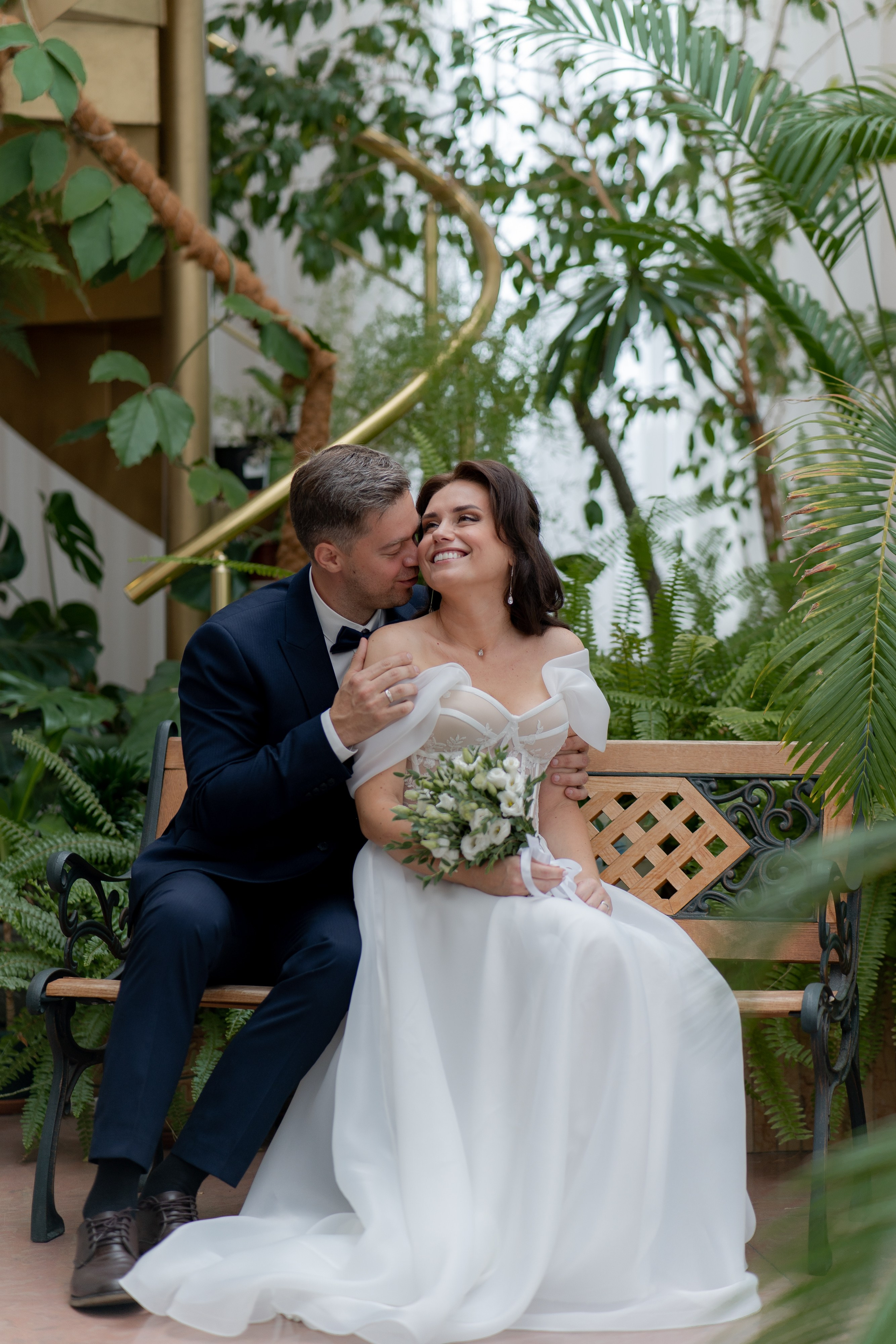 жених целует в ухо невесту в оранжерее загса приморского района