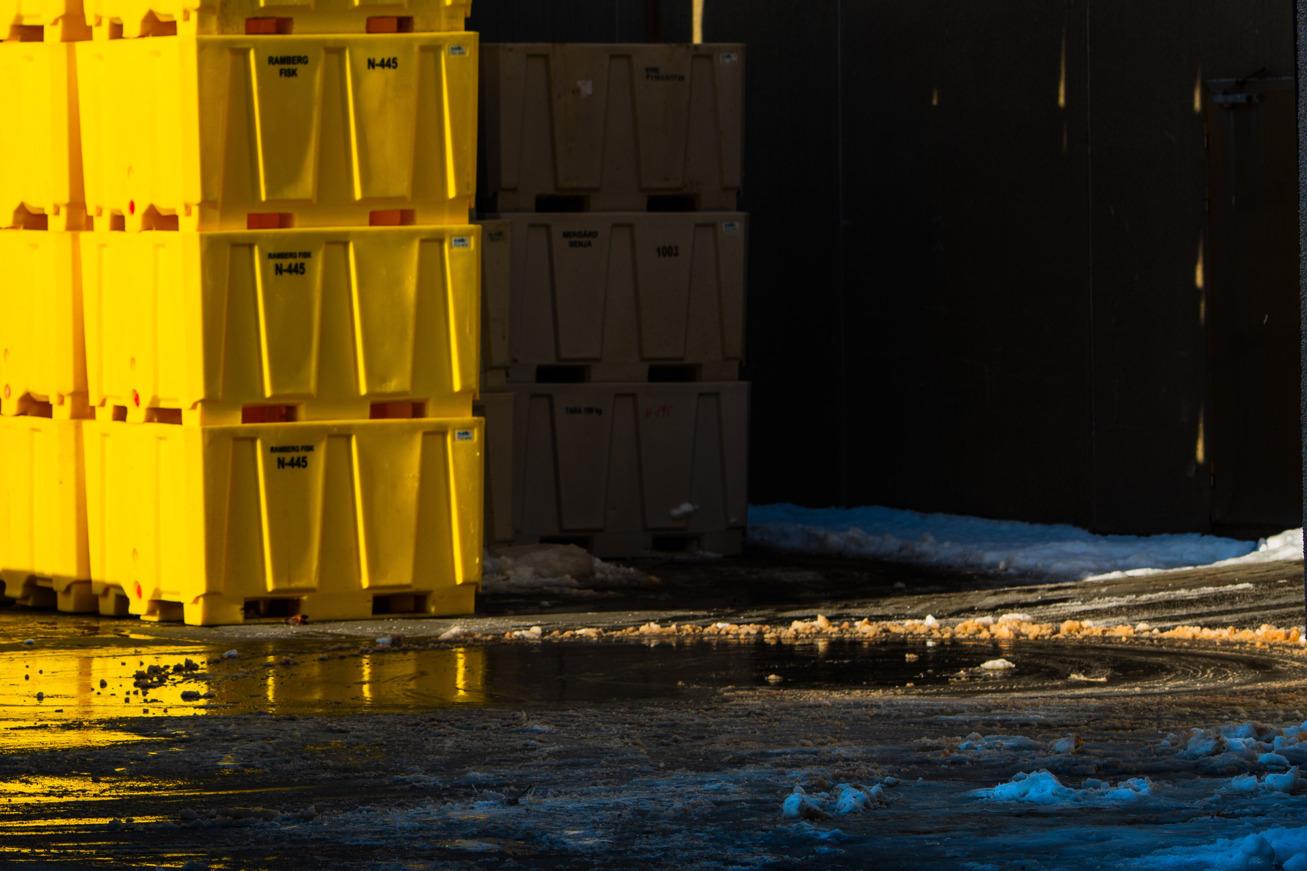Штабели ярко-желтых промышленных пластиковых контейнеров на мокром обледенелом асфальте рядом со снегом и темной стеной здания.