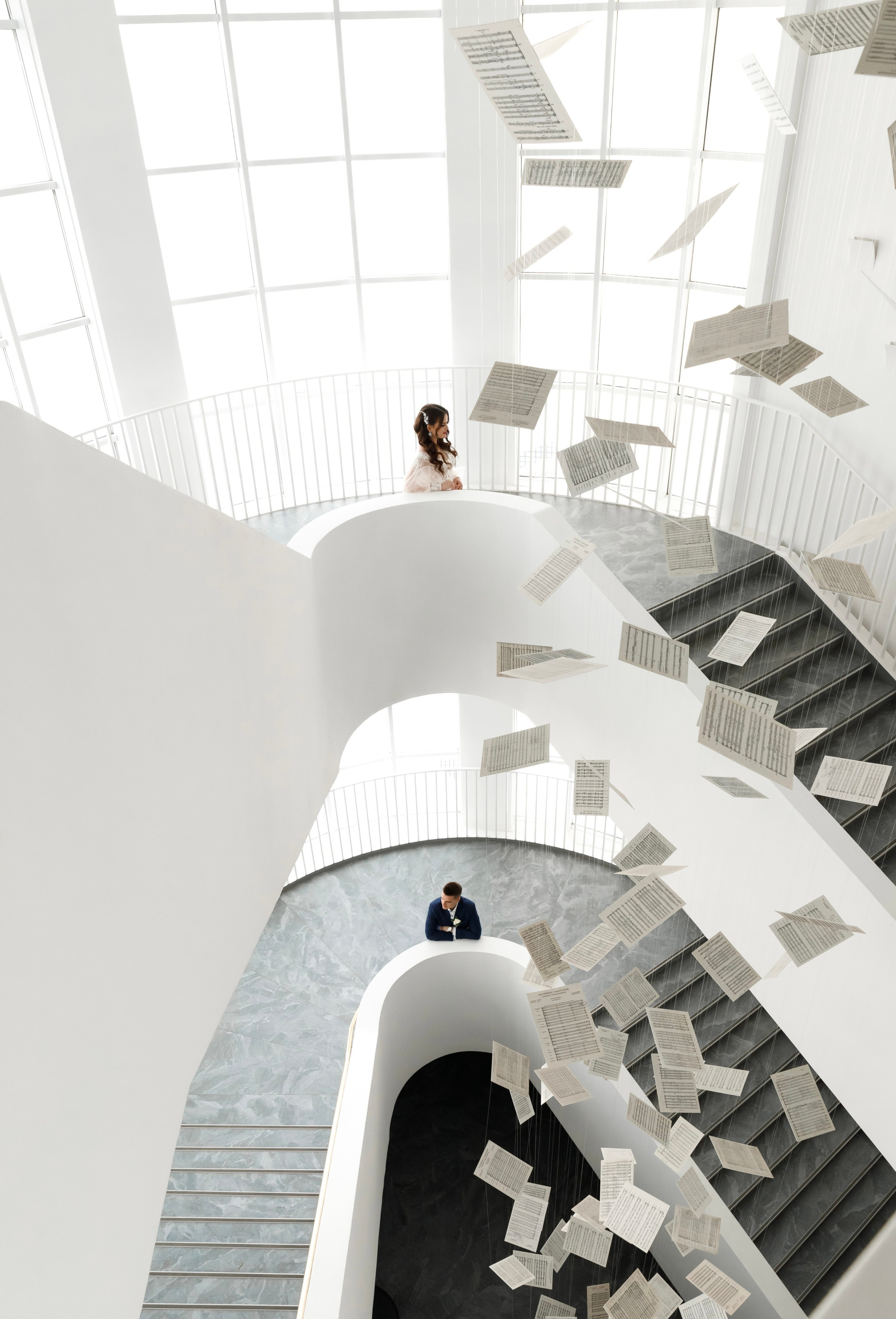 Влюбленные стоят на разных уровнях изогнутой лестницы, ведущей к большому окну. Они смотрят друг на друга с улыбками, возможно, наслаждаясь моментом уединения в этом современном пространстве.