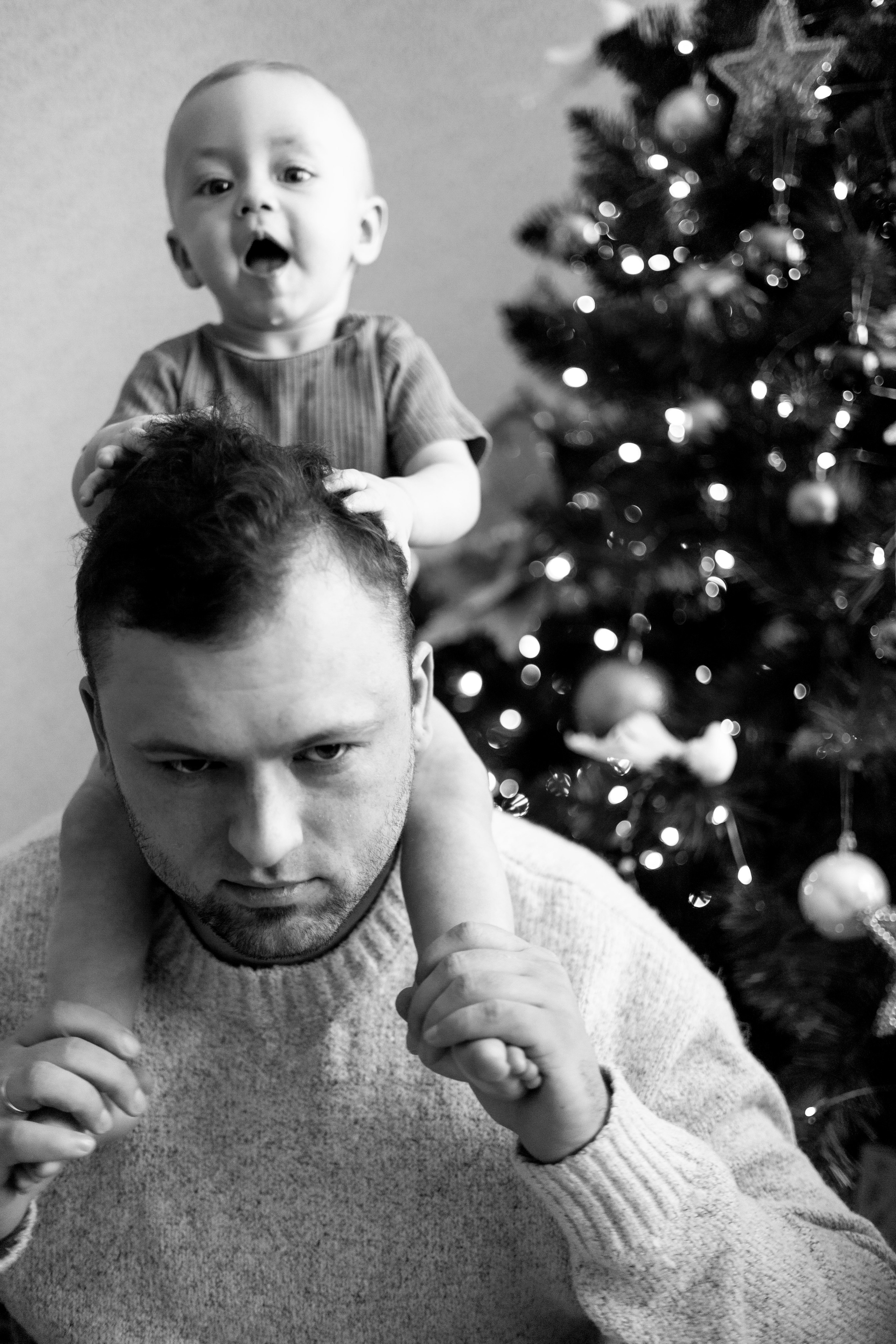 Маленький мальчик сидит на плечах у папы рядом с елкой