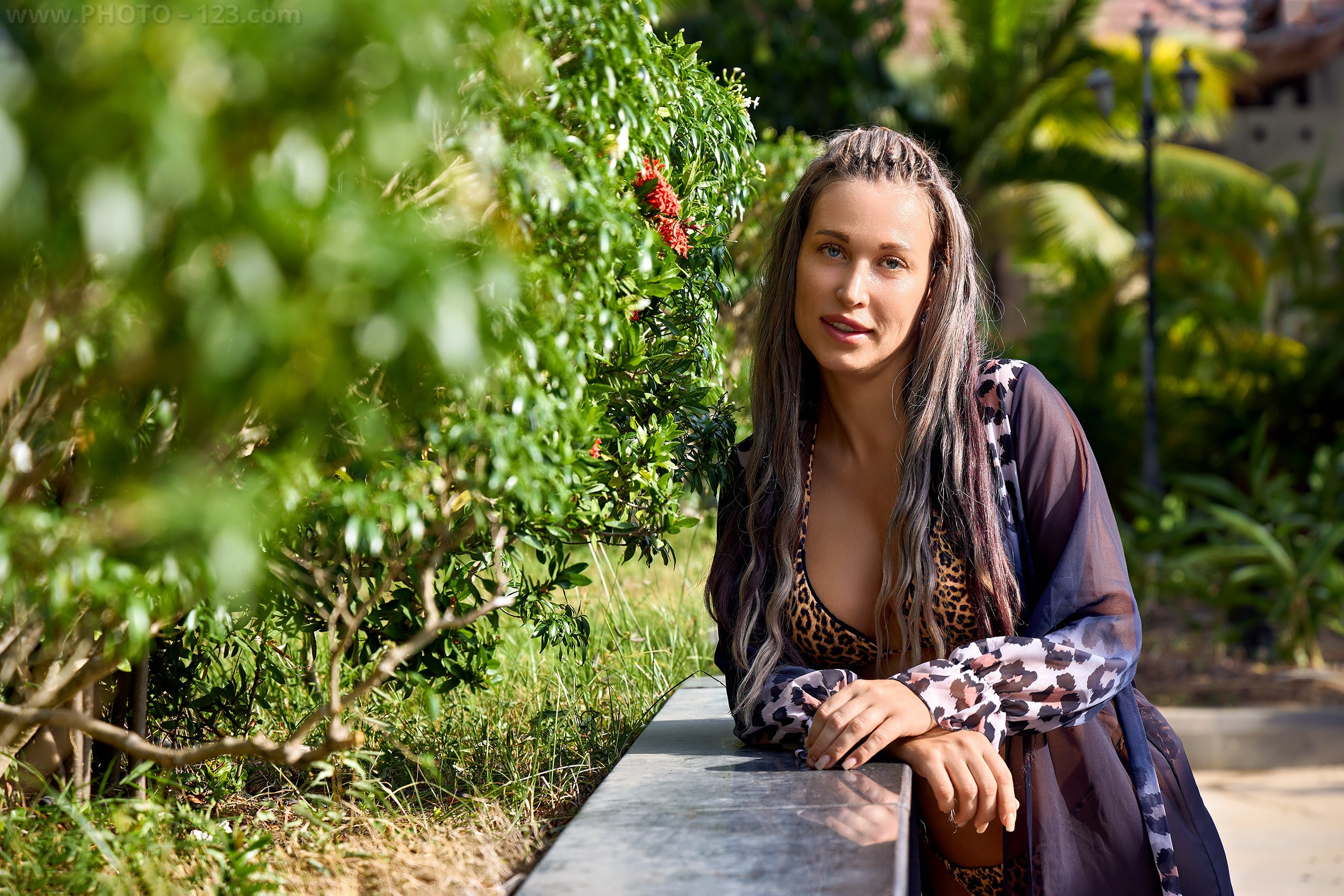 Expressive close-up portrait of a female model leaning on a stone ledge in a vibrant sunny garden setting.