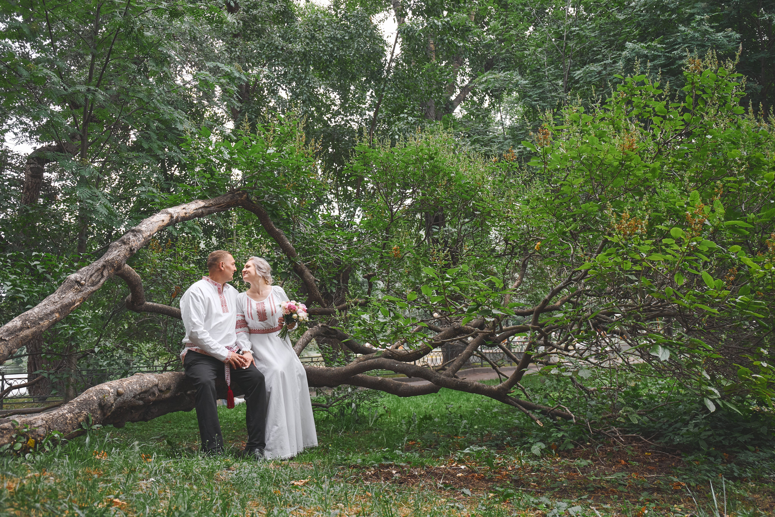 Свадебная пара в традиционных русских нарядах сидят на дереве в парке