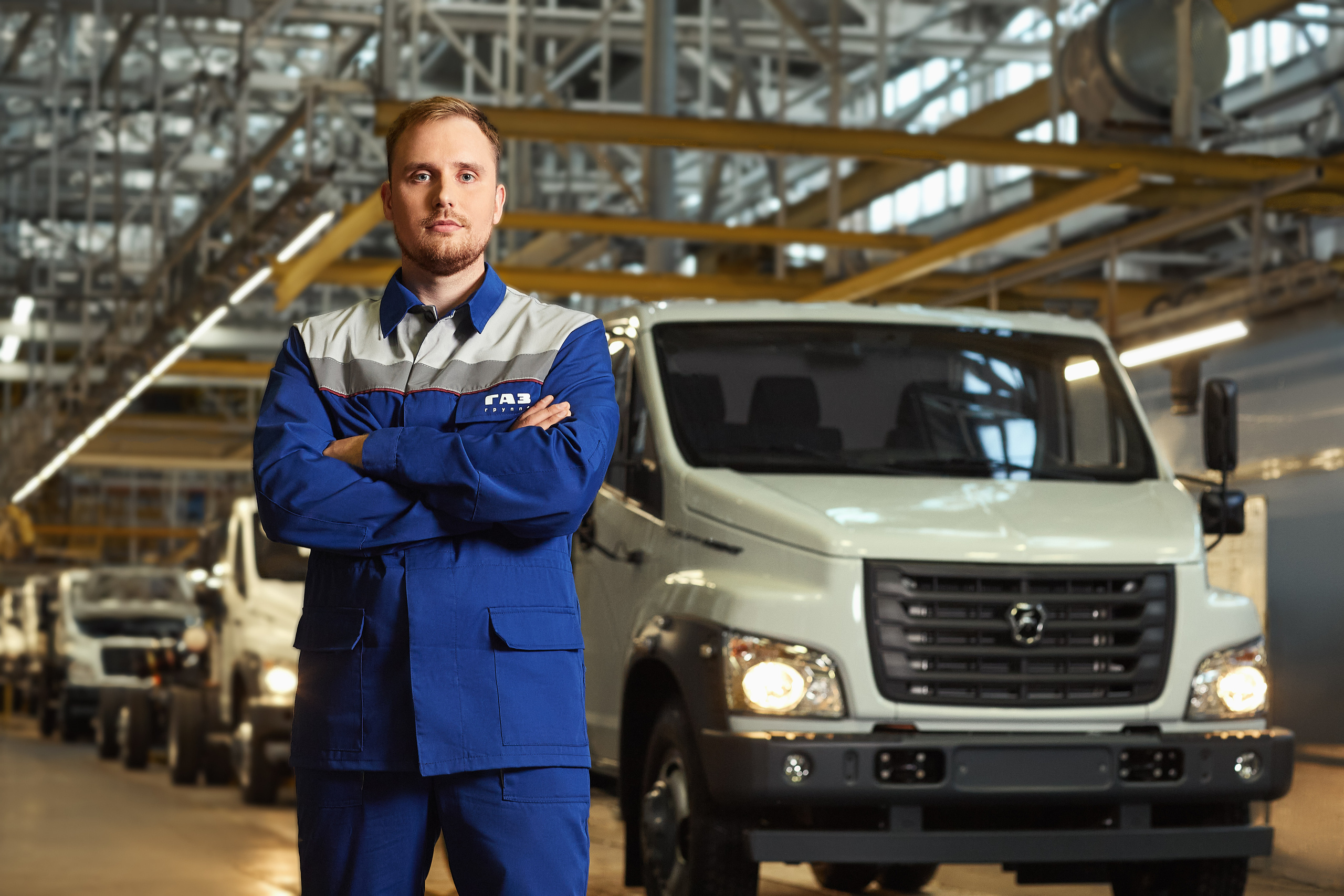 Сотрудник компании гордо стоит сложив руки на груди на фоне автомобильного конвейера.