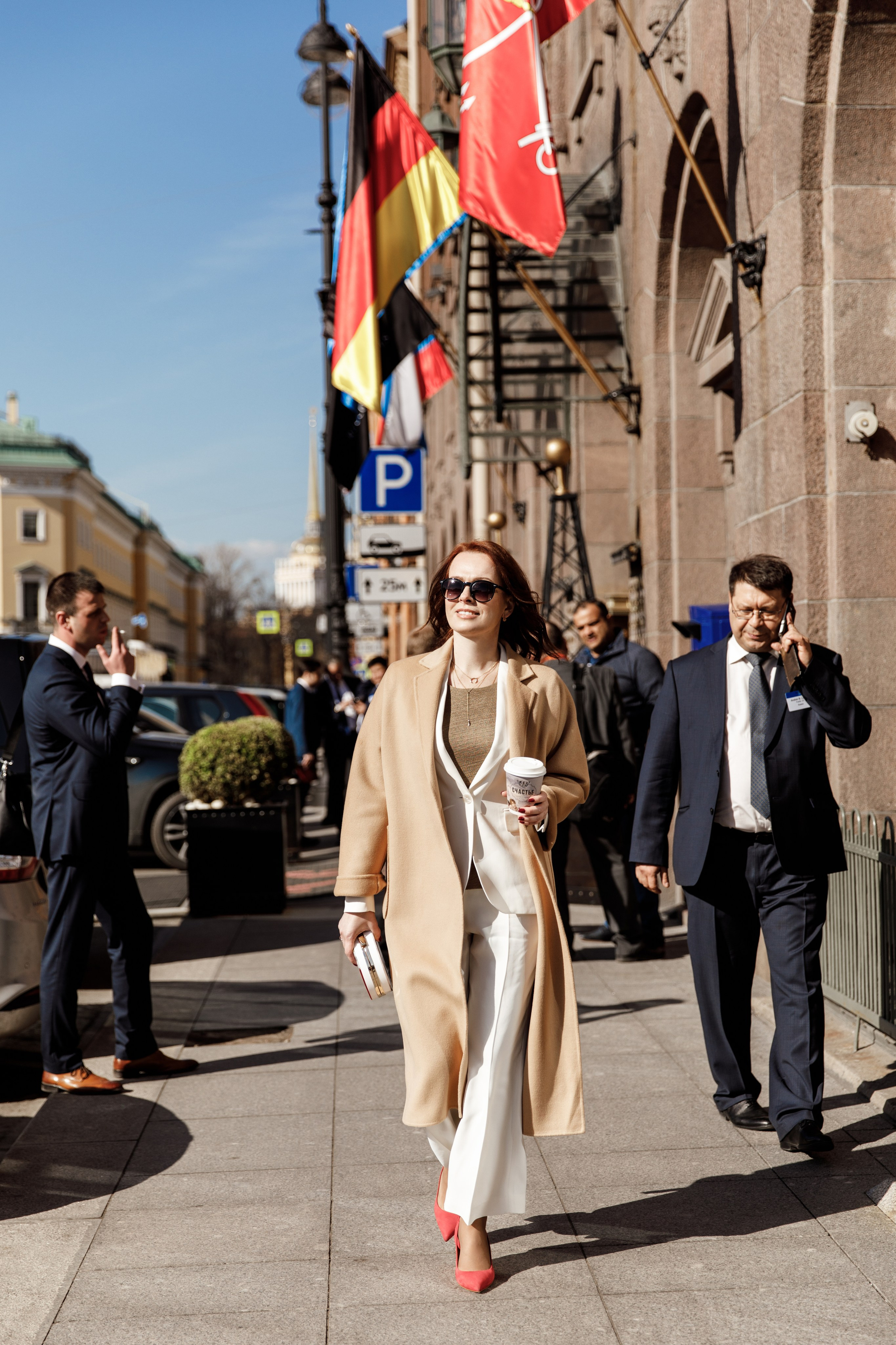 Деловая женщина в бежевом пальто и белых брюках с кофе в руках идет по улице в Санкт-Петербурге