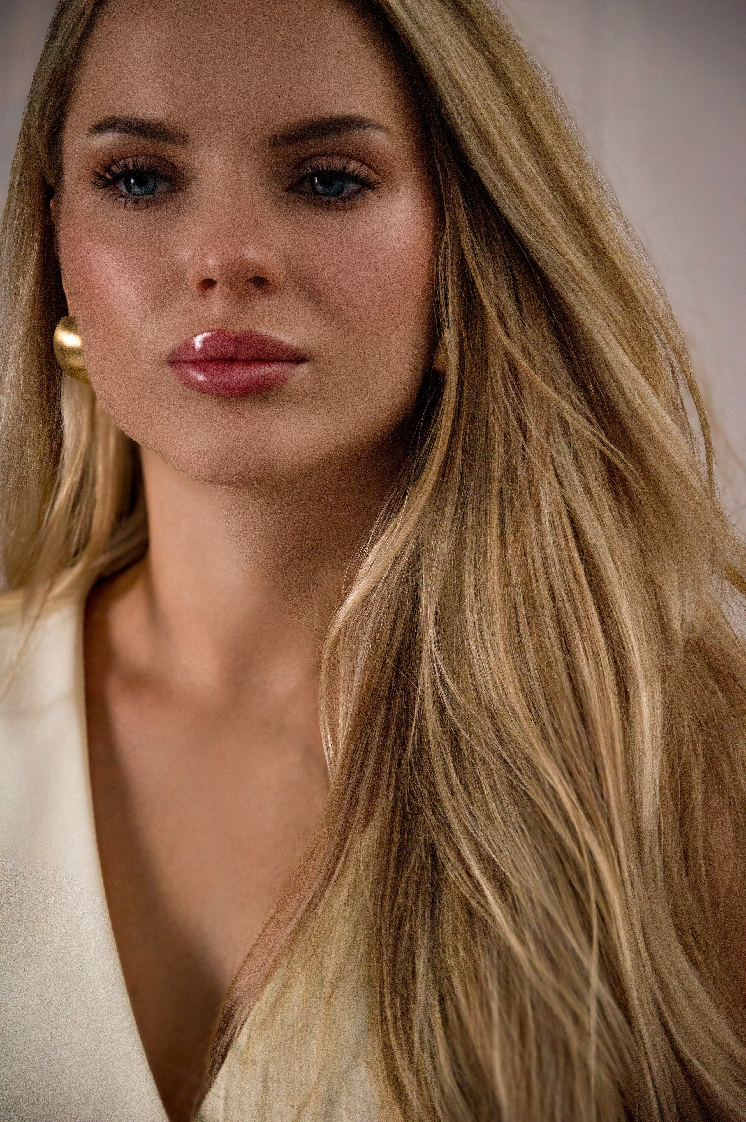 Женщина с длинными светлыми волосами, голубыми глазами, в кремовом платье без рукавов, с крупными круглыми золотыми серьгами, лёгкий макияж, взгляд в сторону.
