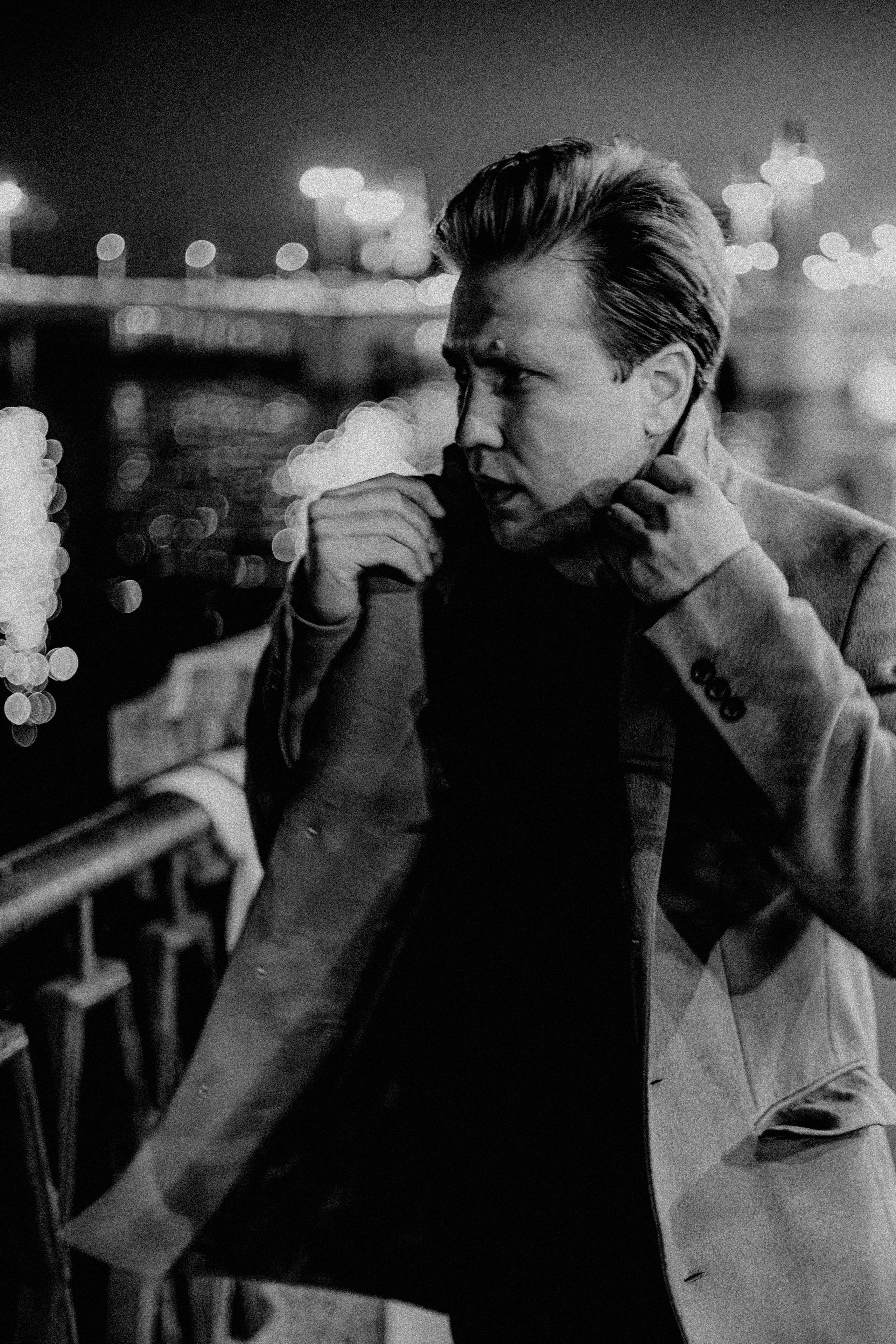 Молодой человек в черной футболке надевает пальто и идет по набережной.