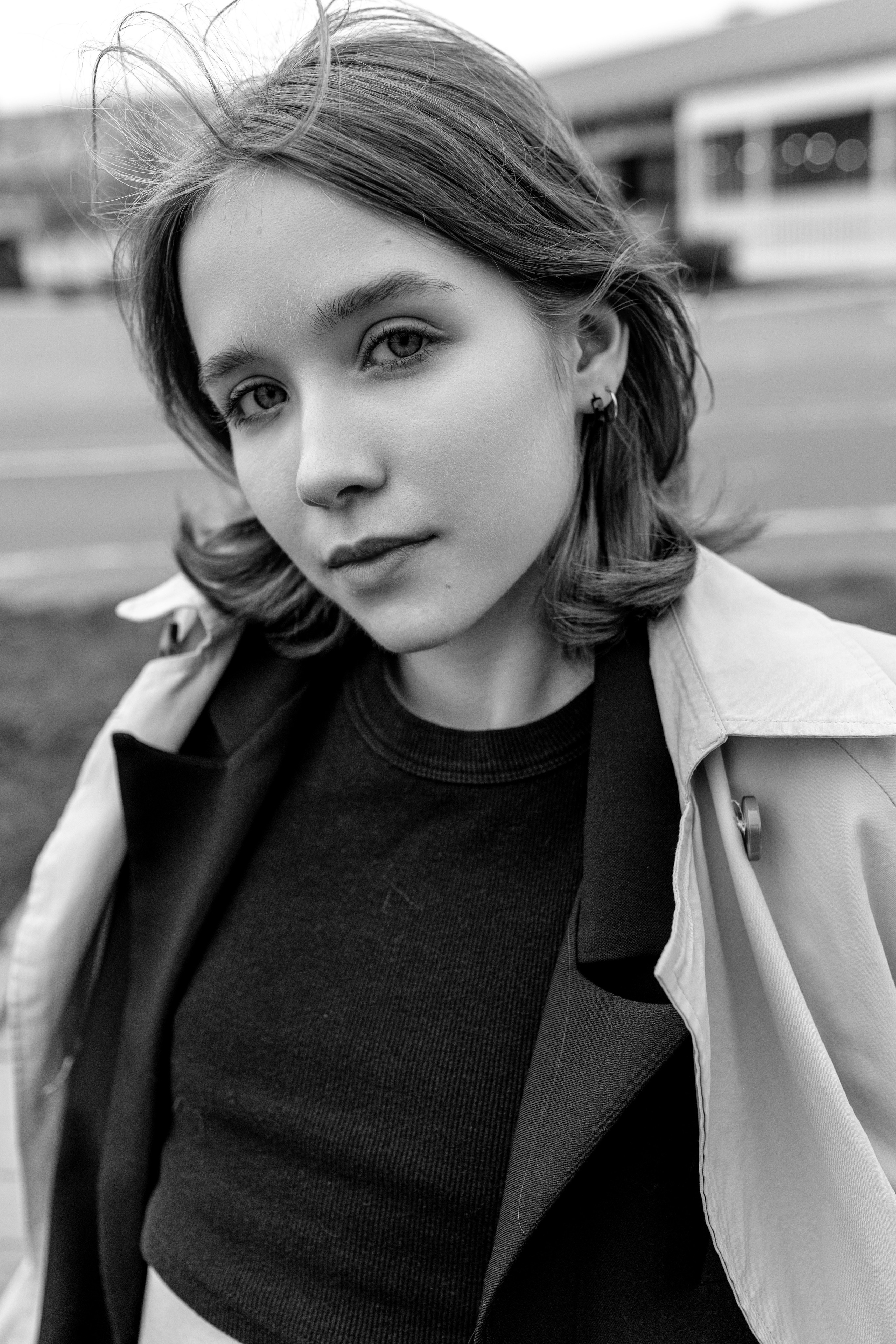 Черно-белый портрет молодой женщины с короткими волосами, одетой в пальто и темный топ, стоящей на улице с уверенным и безмятежным выражением лица.
