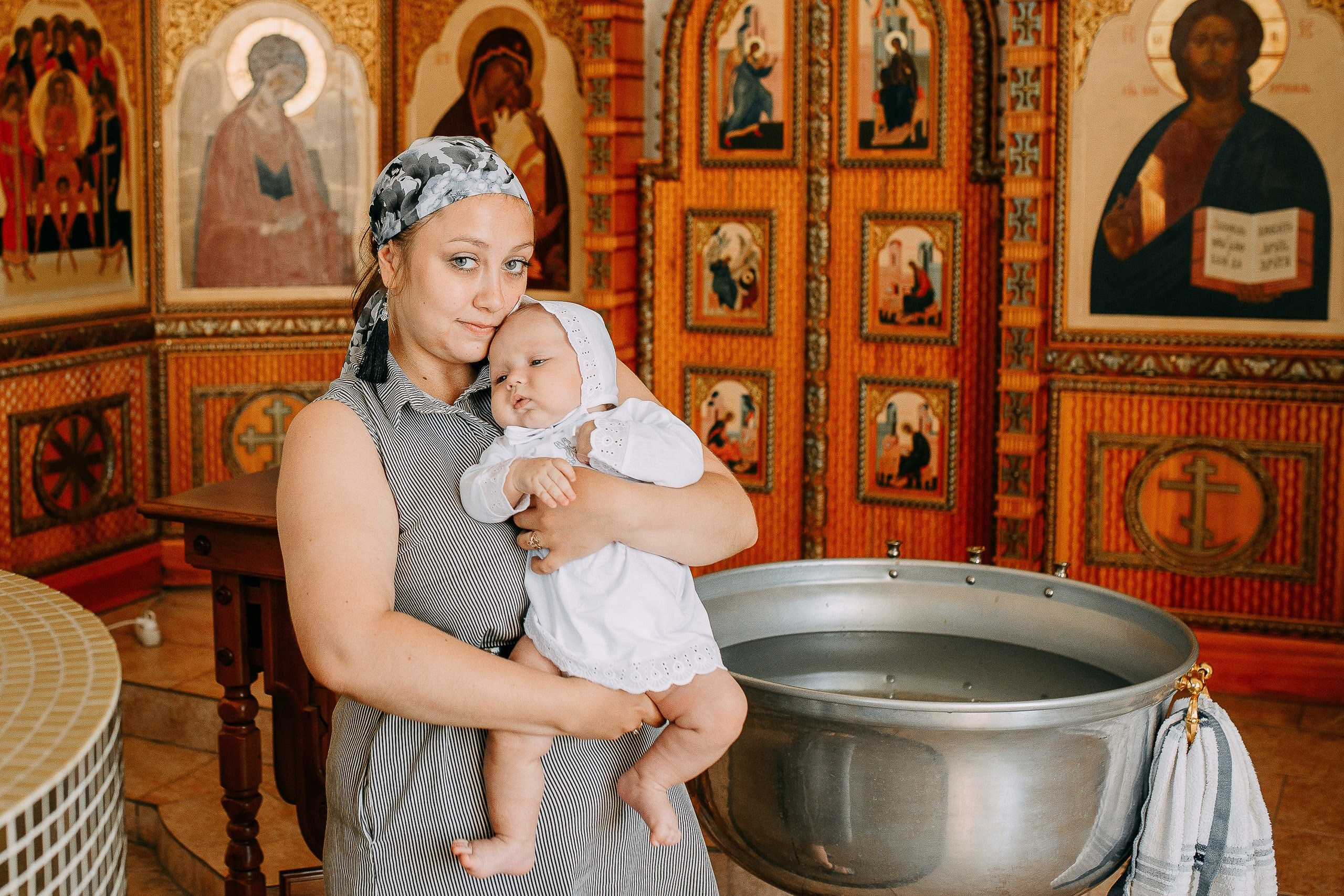 Фотограф на крещение: крестная мама держит младенца в крестильной рубашке перед купелью в храме.