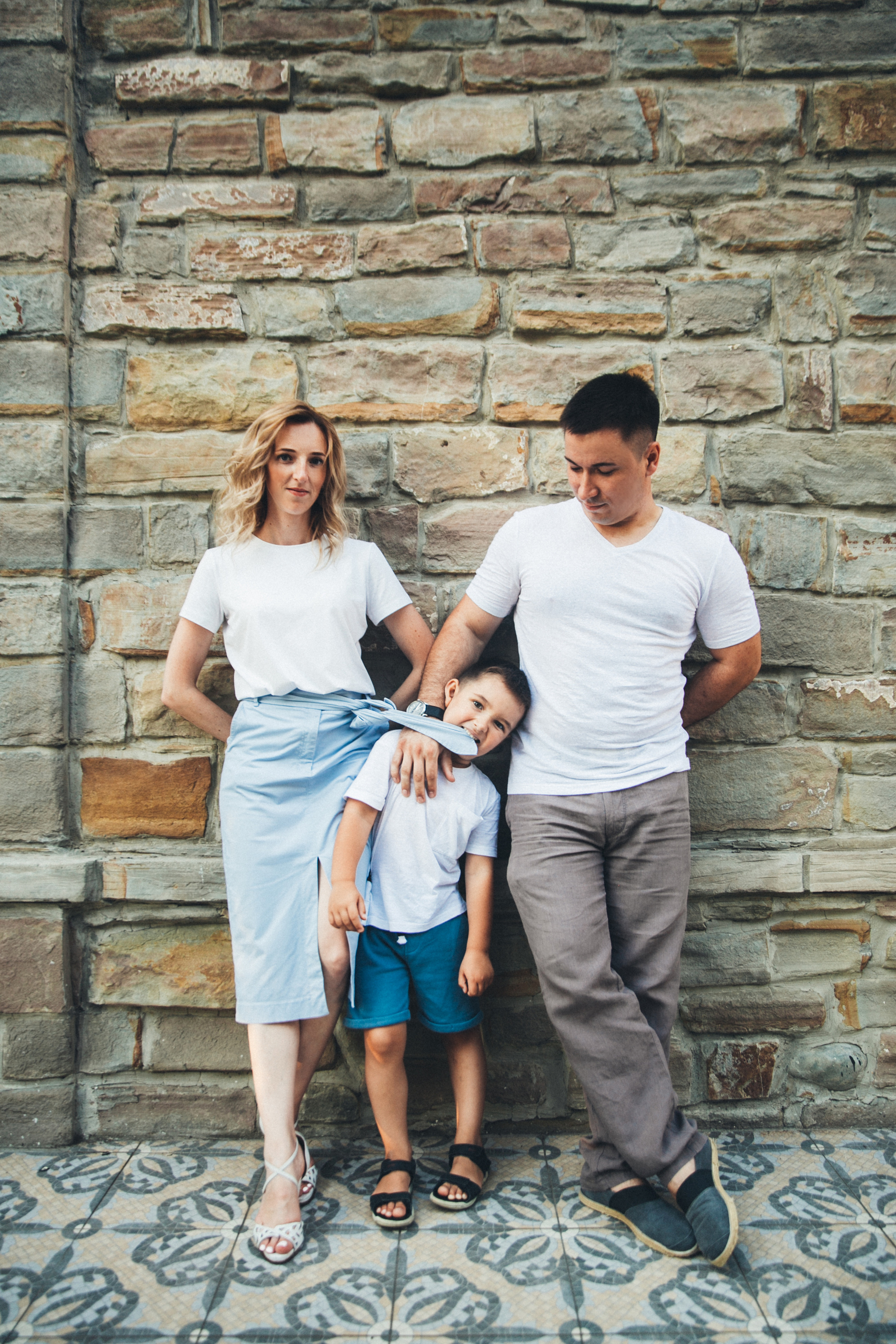 Мама и папа стоят на фоне кирпичной стены а маленький сын кусает мамин пояс от юбки