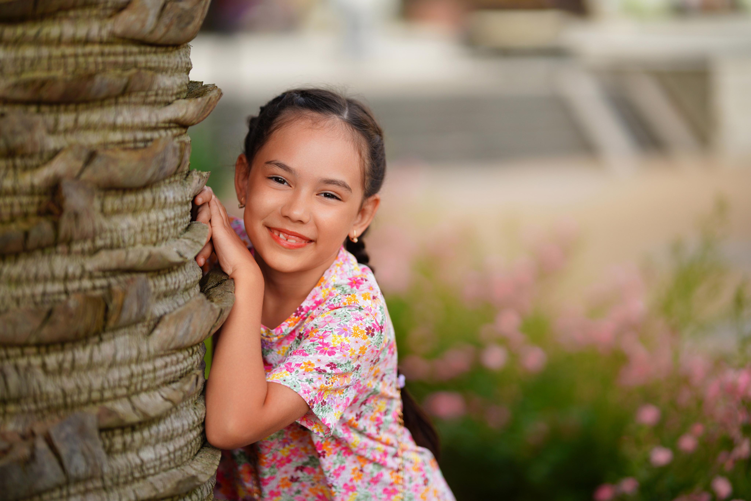 Маленькая девочка улыбается в камеру облокотившись на ствол пальмы