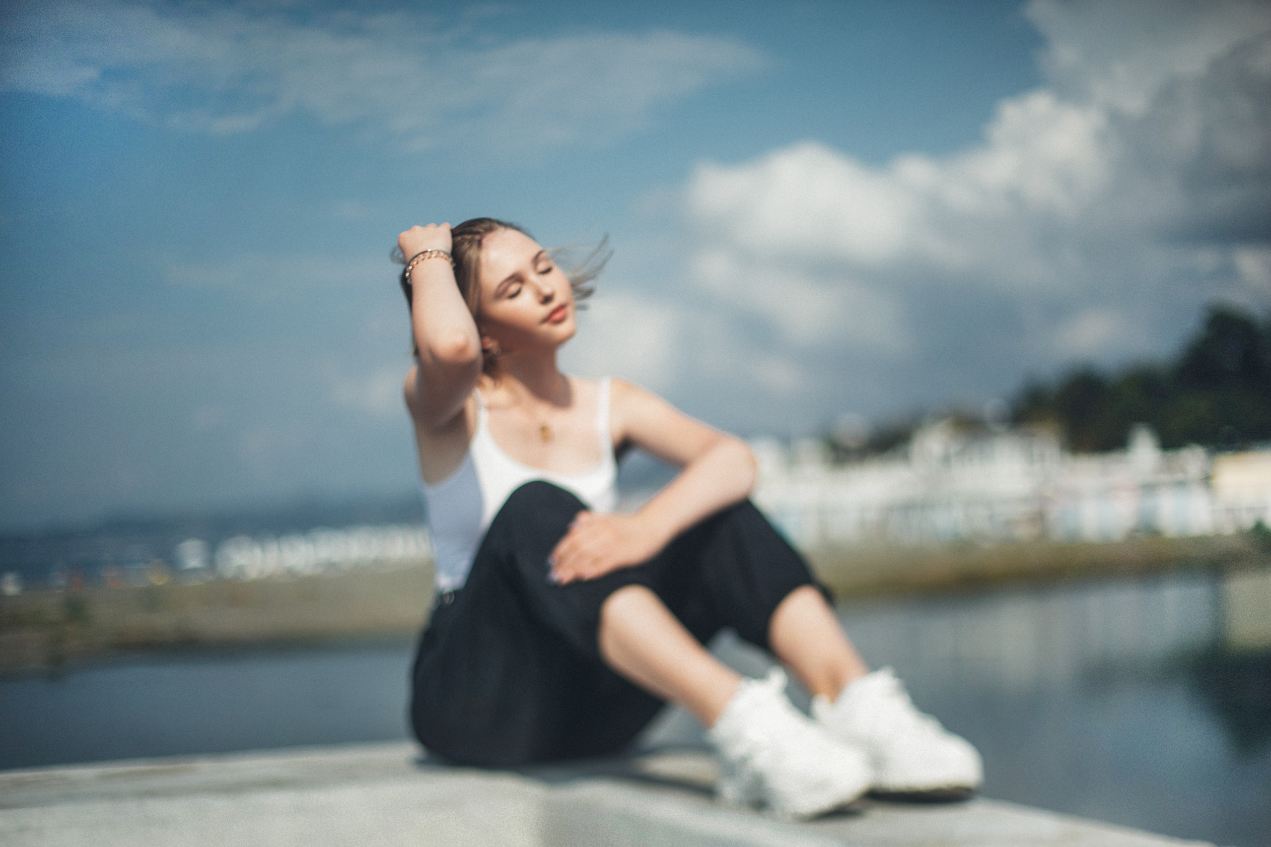 Девушка сидит на бордюре и поправляет свои волосы на фоне реки и облачного неба