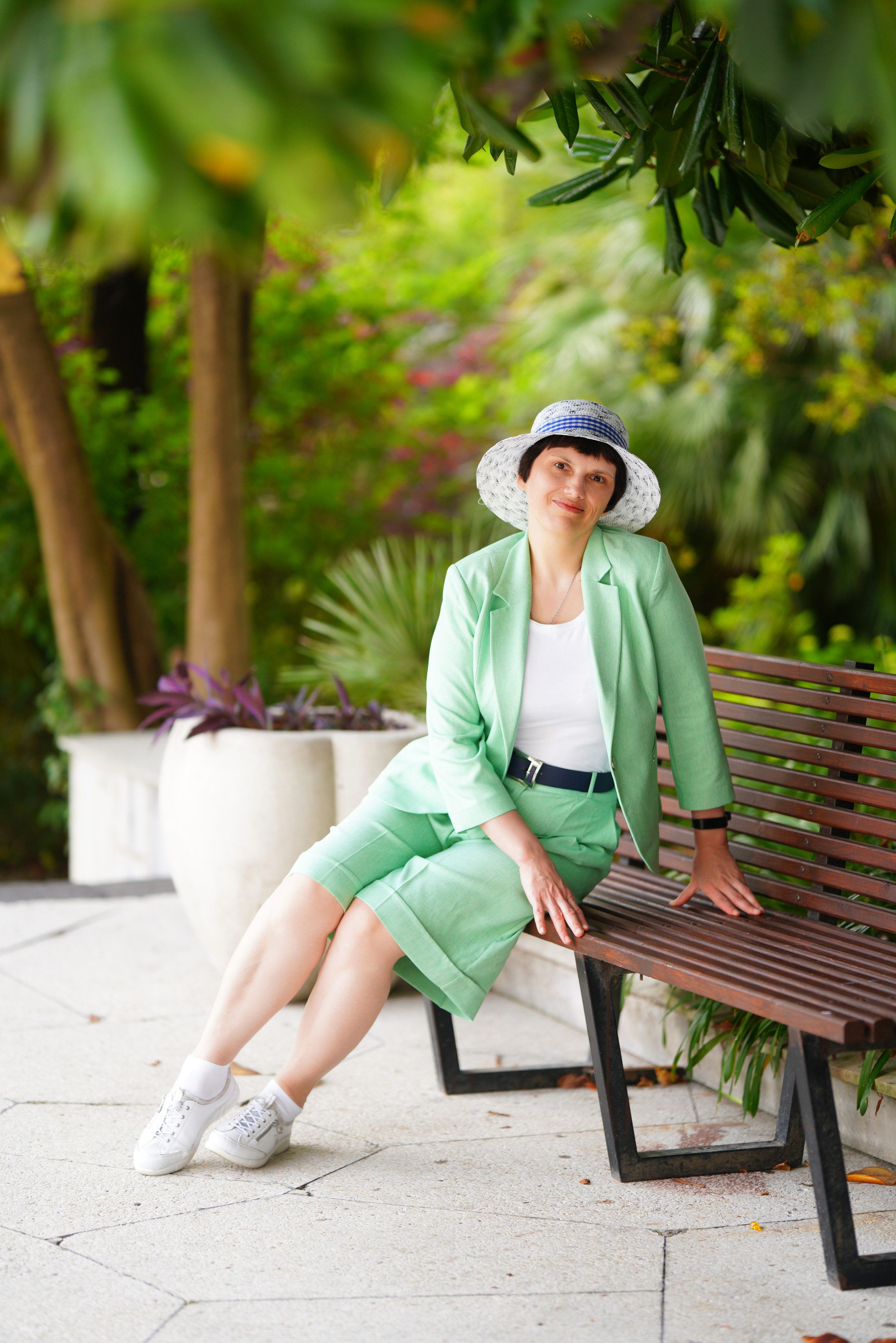 Элегантная женщина в зелёном костюме и шляпе сидит на лавочке среди зелени