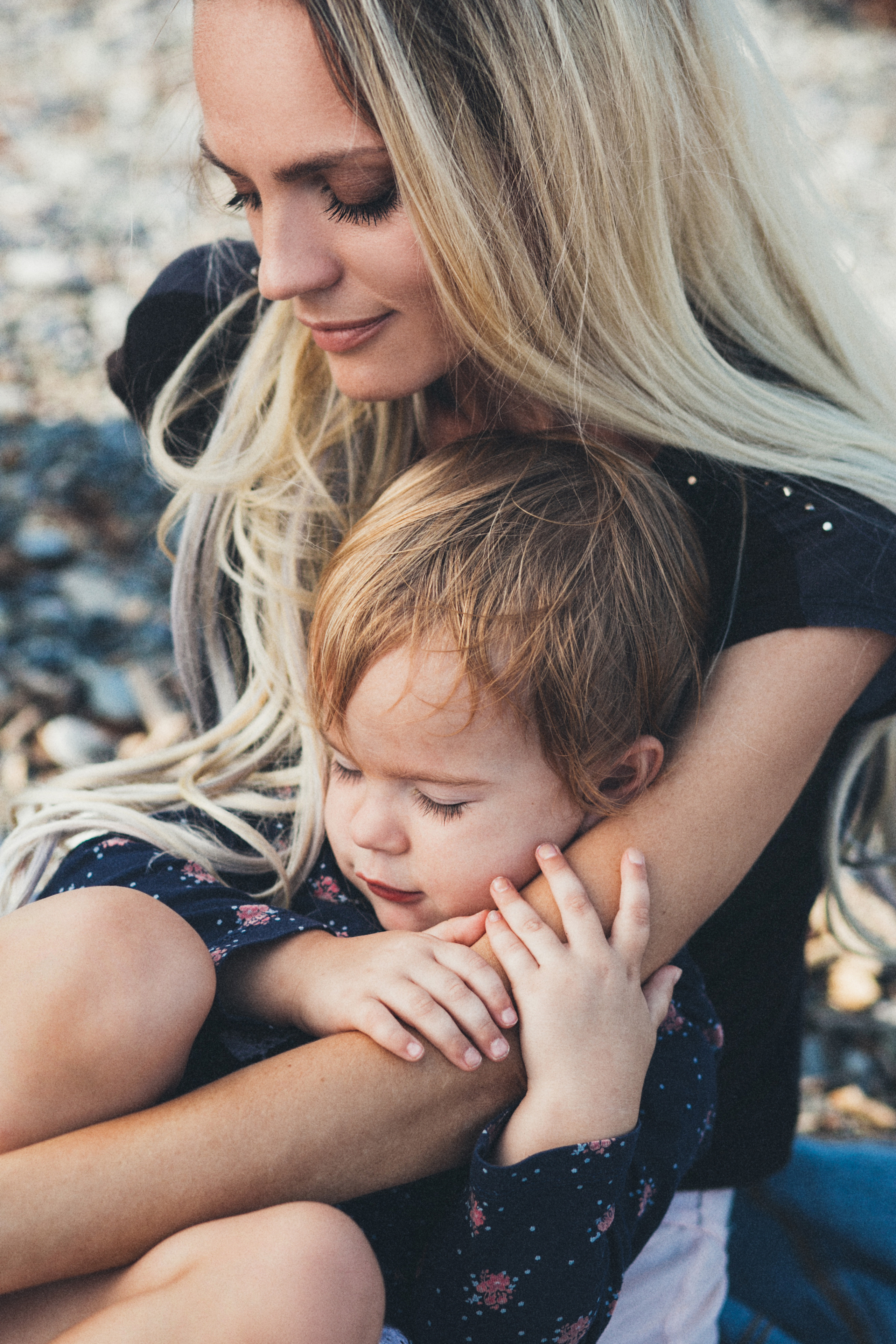 Мама обнимая, держит на руках дочь, сидя на гальке