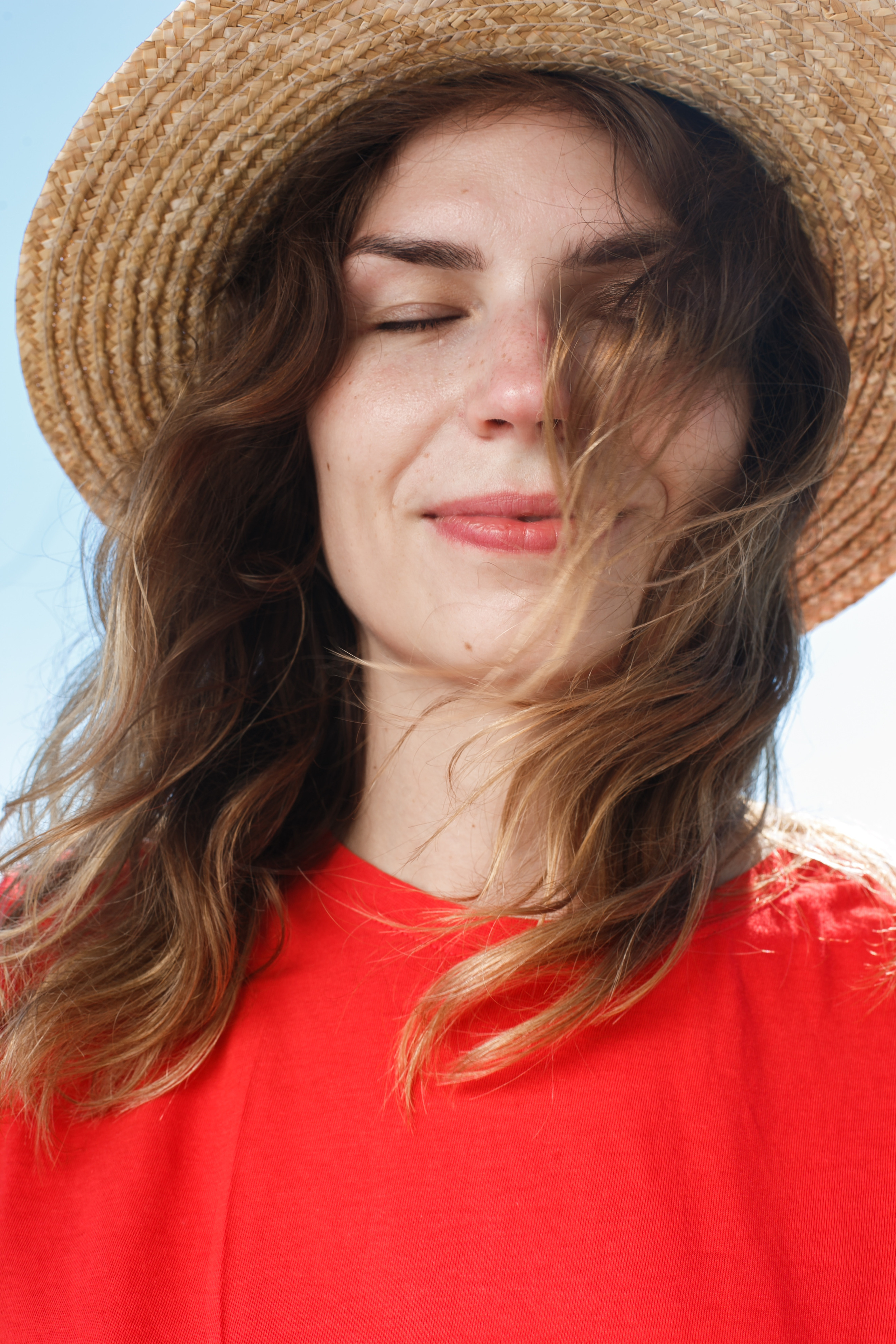 Женщина в шляпе и в красной футболке улыбается закрытыми глазами, а её волосы развивает ветер