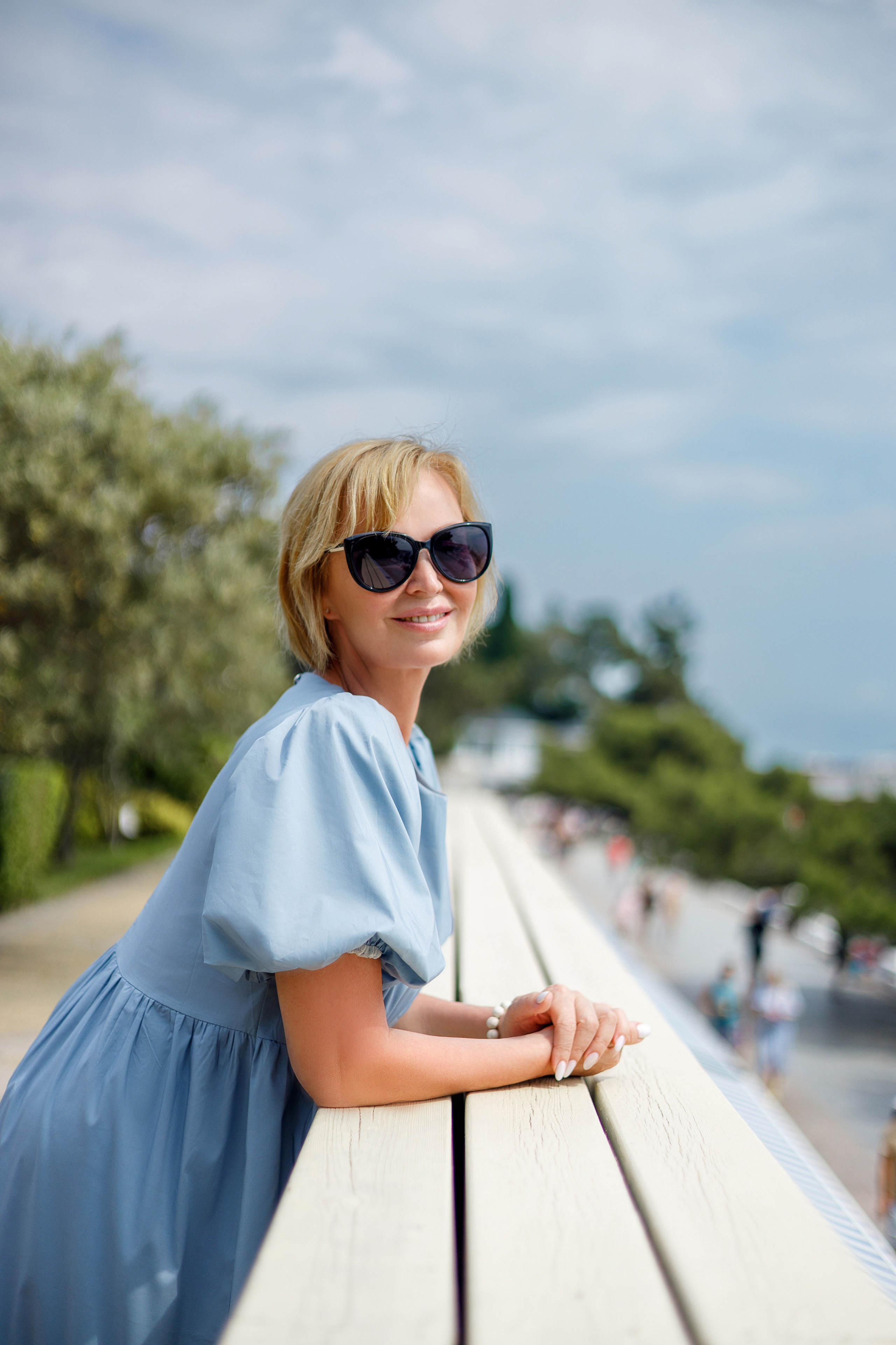Взрослая женщина с белыми волосами, в очках, и голубом платье стоит облокотившись на бордюр на фоне голубого неба