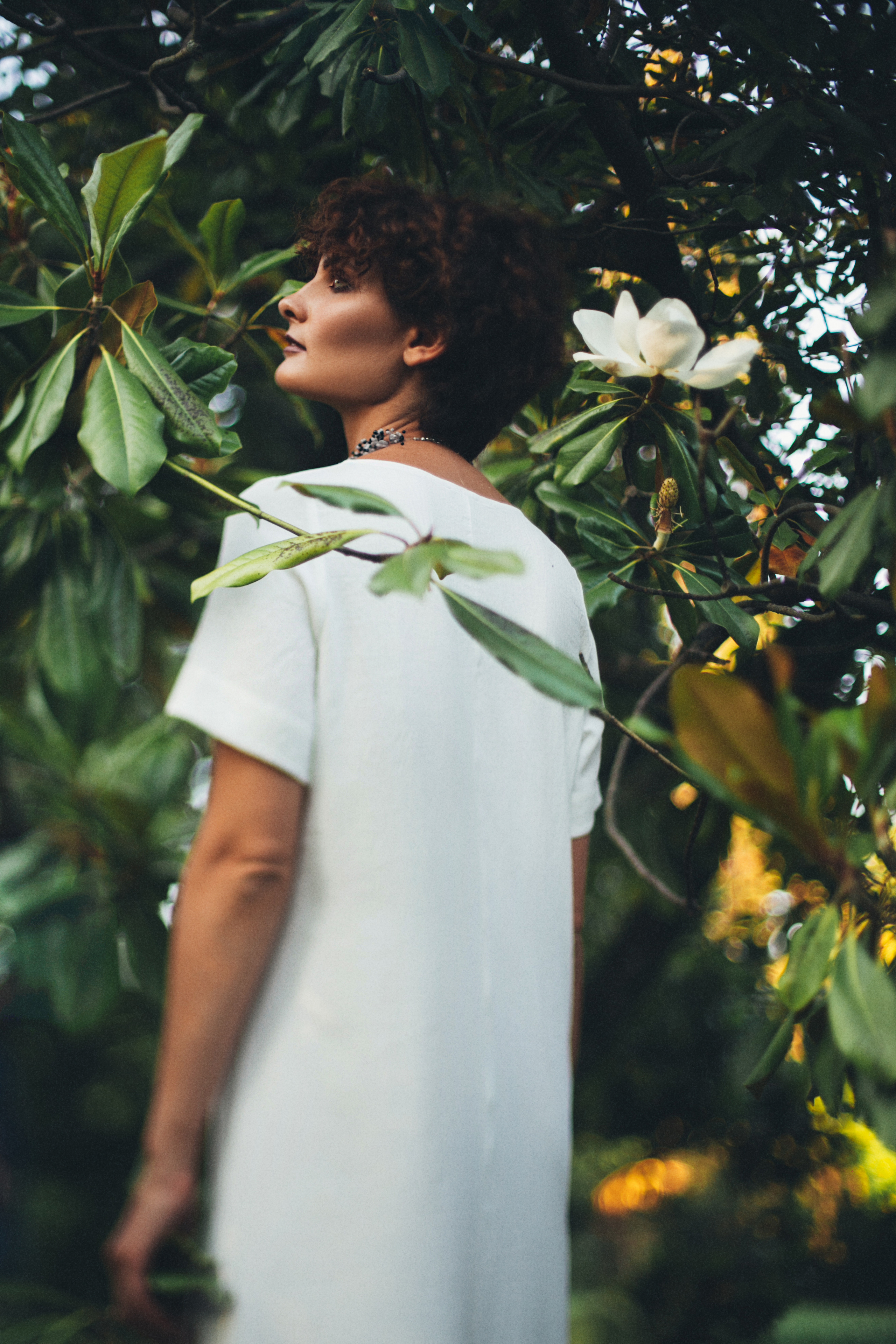 Девушка стоит спиной повернув голову набок в дереве магнолии рядом с её головой цветок