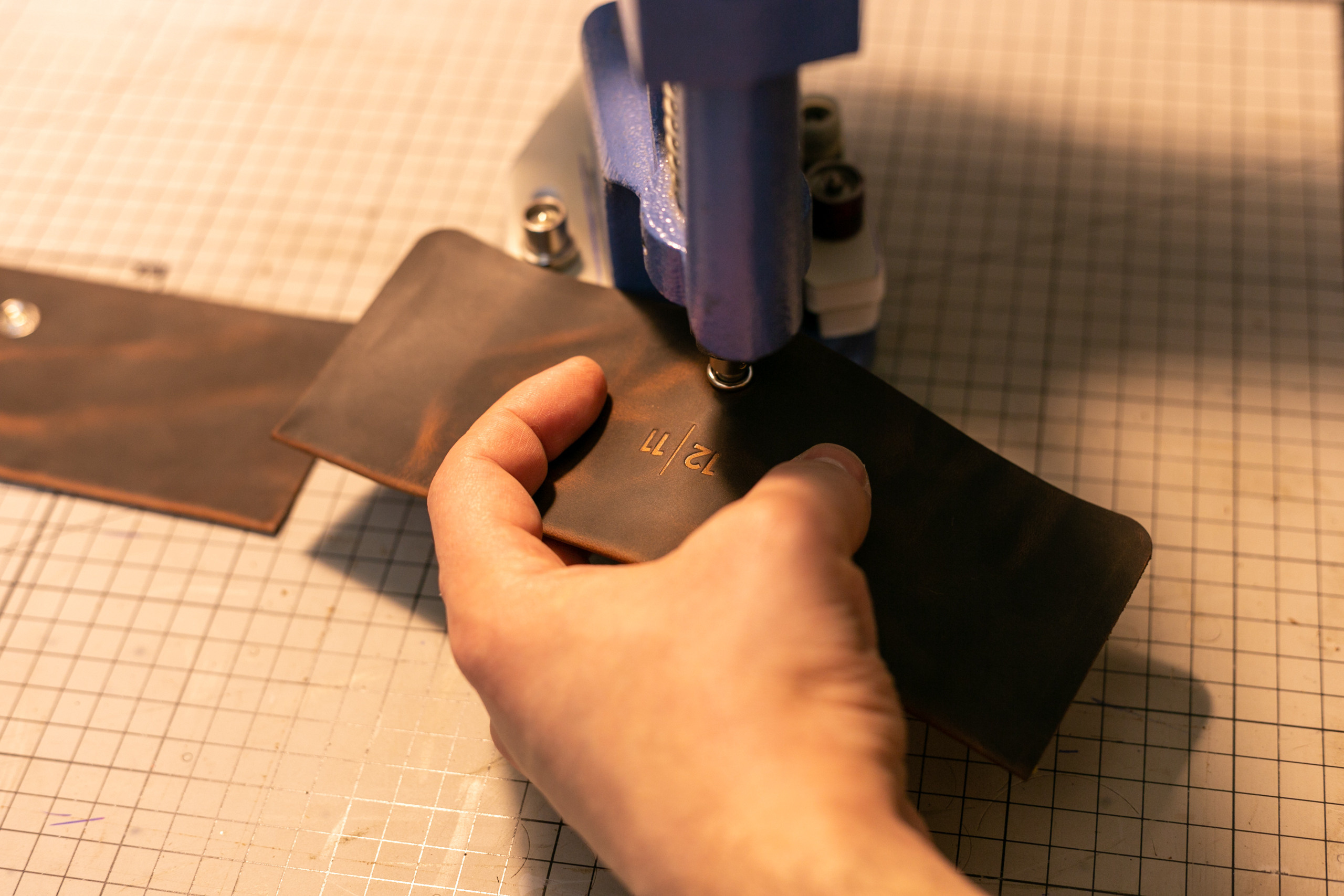 Руки крупно держат кусок коричневой кожи и ставят на него кнопку дыроколом. Машинка для установки кнопок.