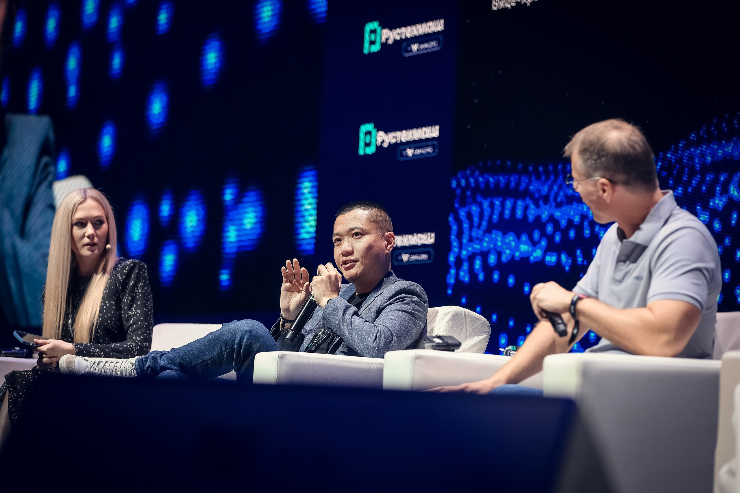 Выступление представителя криптобиржи BINGX на Blockchain 2025 в MTS Hall, Москва. Три спикера на сцене, акцент на докладчике с микрофоном.