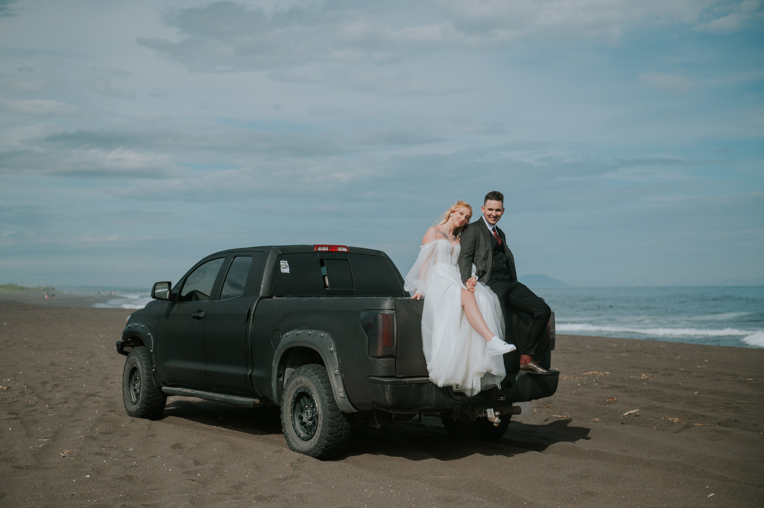 Жених в костюме и невеста в белом платье сидят в кузове чёрного пикапа на чёрном пляже у моря. Атмосфера расслабленная, романтичная.