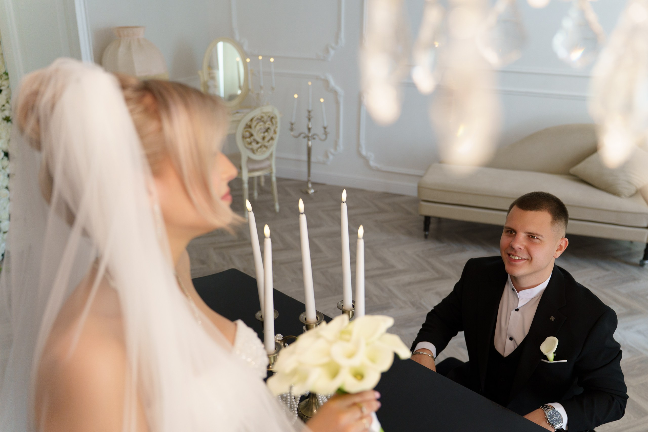 Жених в классическом чёрном костюме с бутоньеркой сидит за столом и с улыбкой смотрит на невесту. Невеста в белом свадебном платье с фатой держит букет белых калл. На столе зажжены свечи, интерьер украшен люстрой и элегантной мебелью | свадебные фотографии с эмоциями Екатеринбург