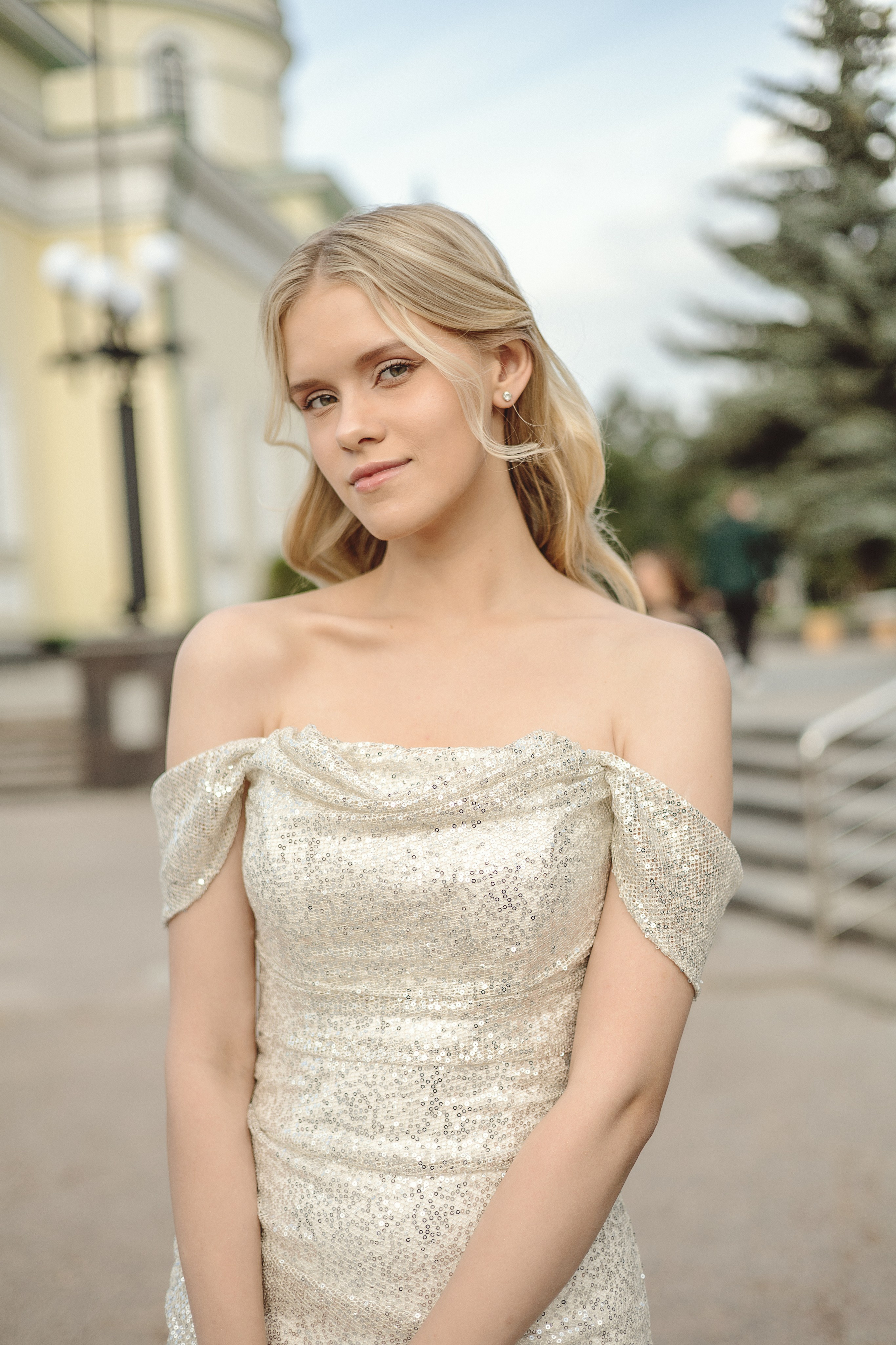 Молодая девушка в элегантном серебристом платье с открытыми плечами стоит на улице. У нее светлые волосы и мягкая улыбка, от нее исходит аура спокойствия и уверенности.