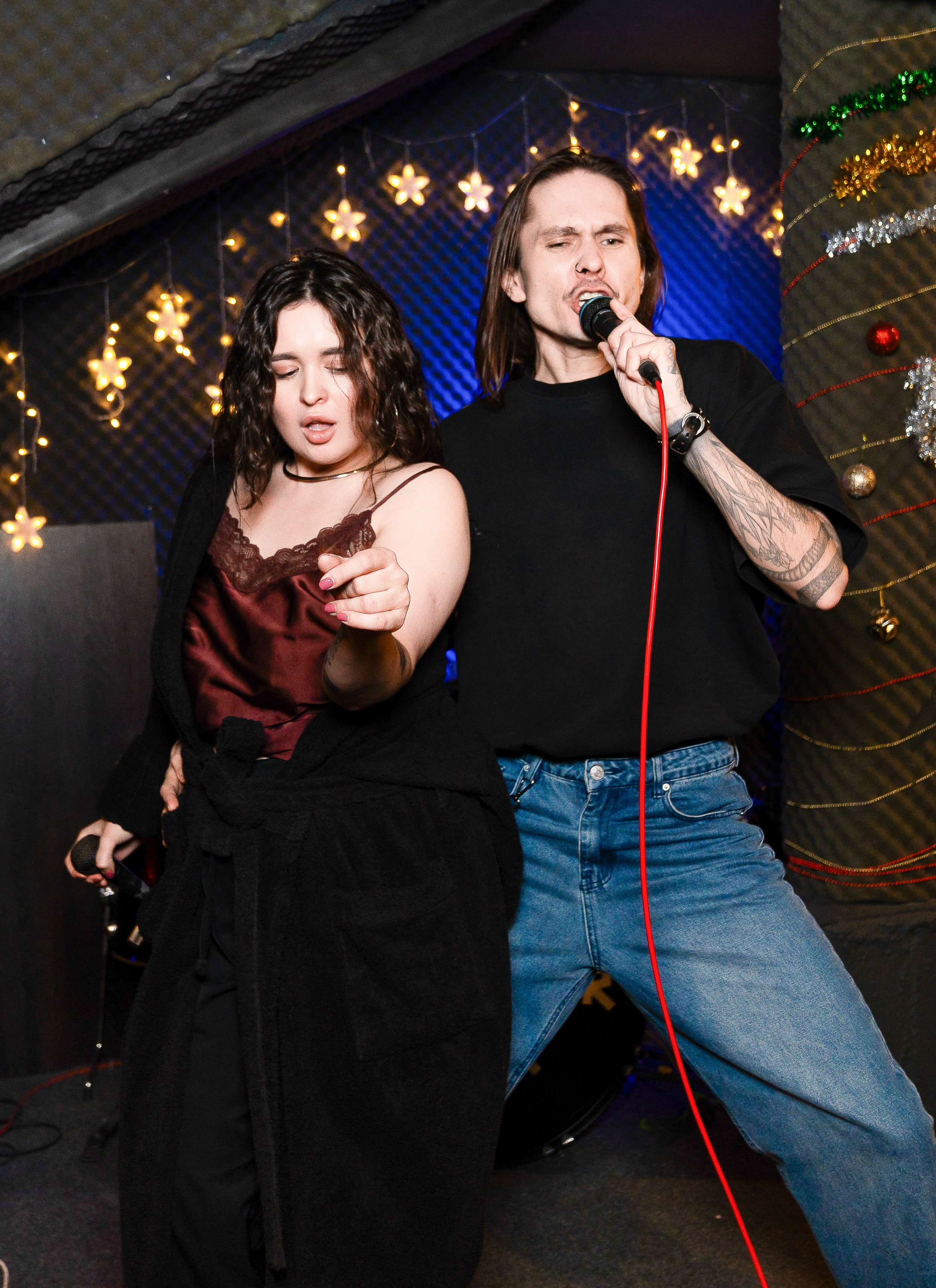 Дуэт на сцене: девушка в бордовом топе и парень с длинными волосами эмоционально поют в микрофон под гирляндами. Живая репортажная съёмка выступления, фотограф Мальцев Евгений.