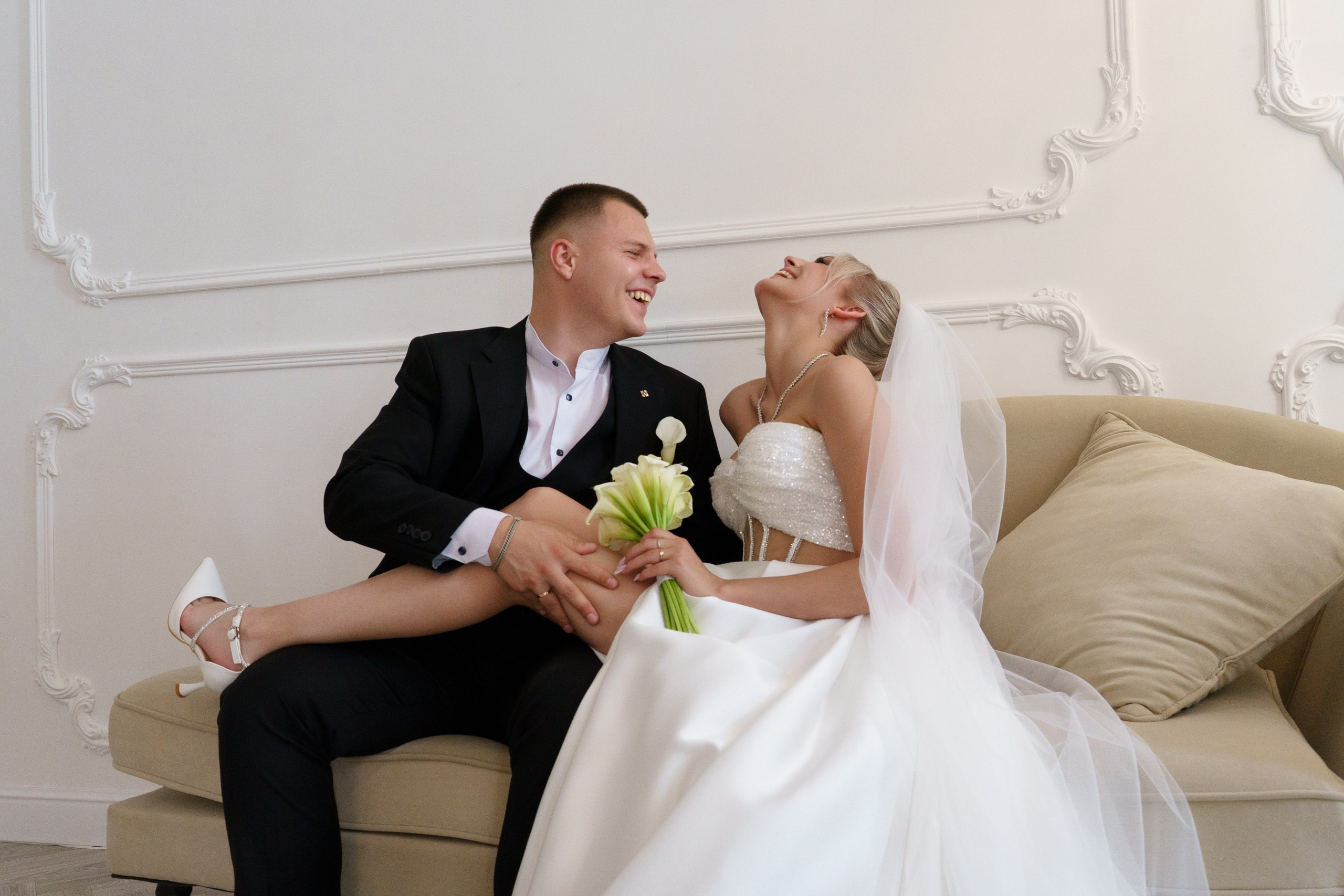 Счастливые молодожёны на свадебной фотосессии: они сидят на диване, невеста в белом платье с фатой держит букет, жених — в чёрном костюме. Оба выглядят влюблёнными и радостными | свадебные фото в студии Екатеринбург