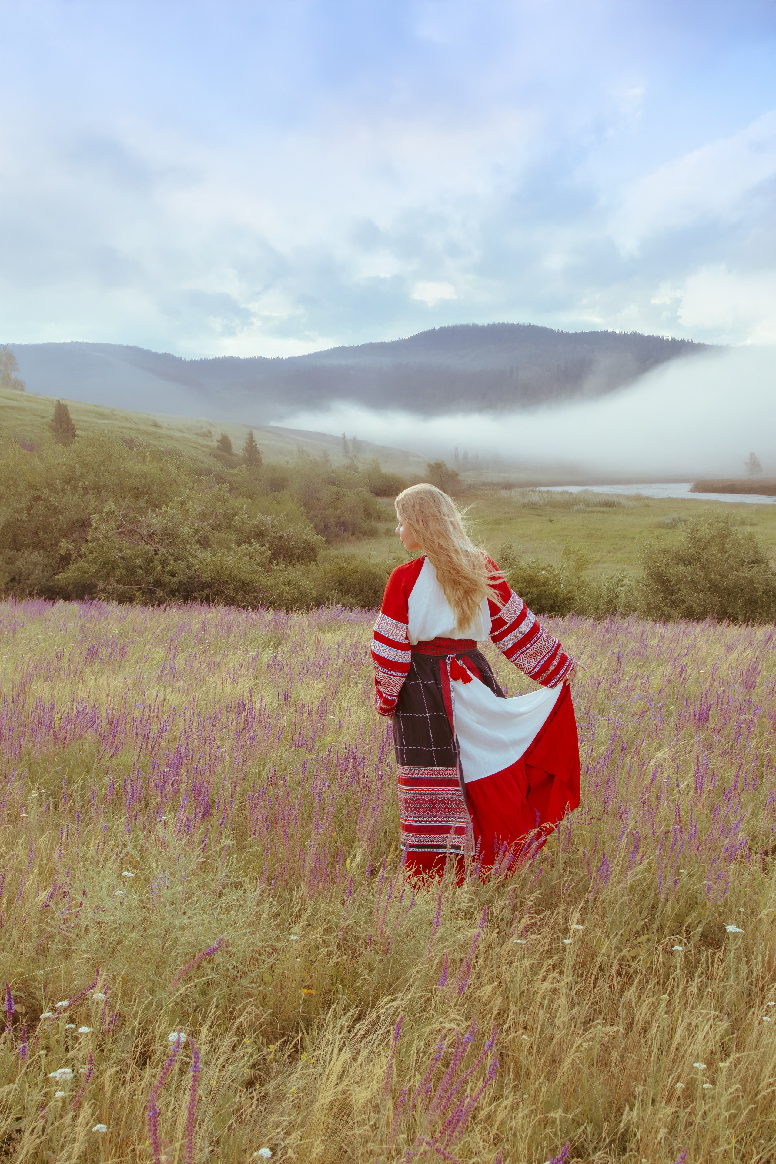Девушка в славянском платье стоит на фоне туманных холмов и красивым небом