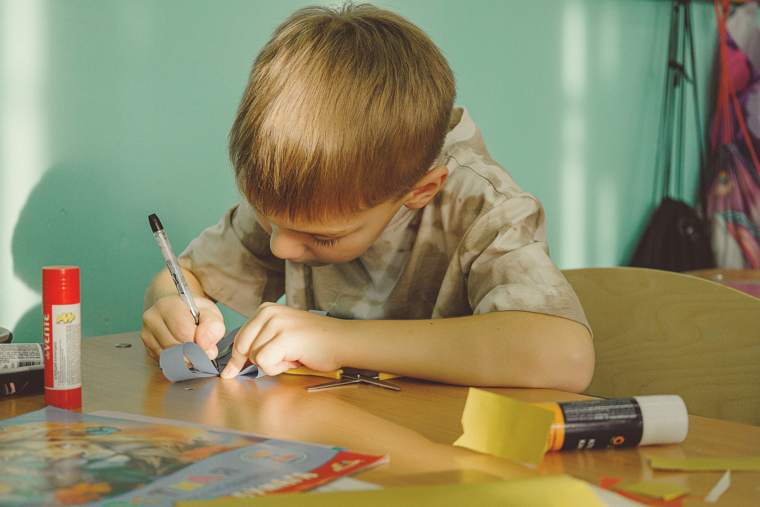 Ребенок со светло-каштановыми волосами сосредоточенно рисует за деревянной партой в школе. В лучах теплого солнечного света его окружают такие художественные принадлежности, как клей, ножницы и разноцветная бумага.