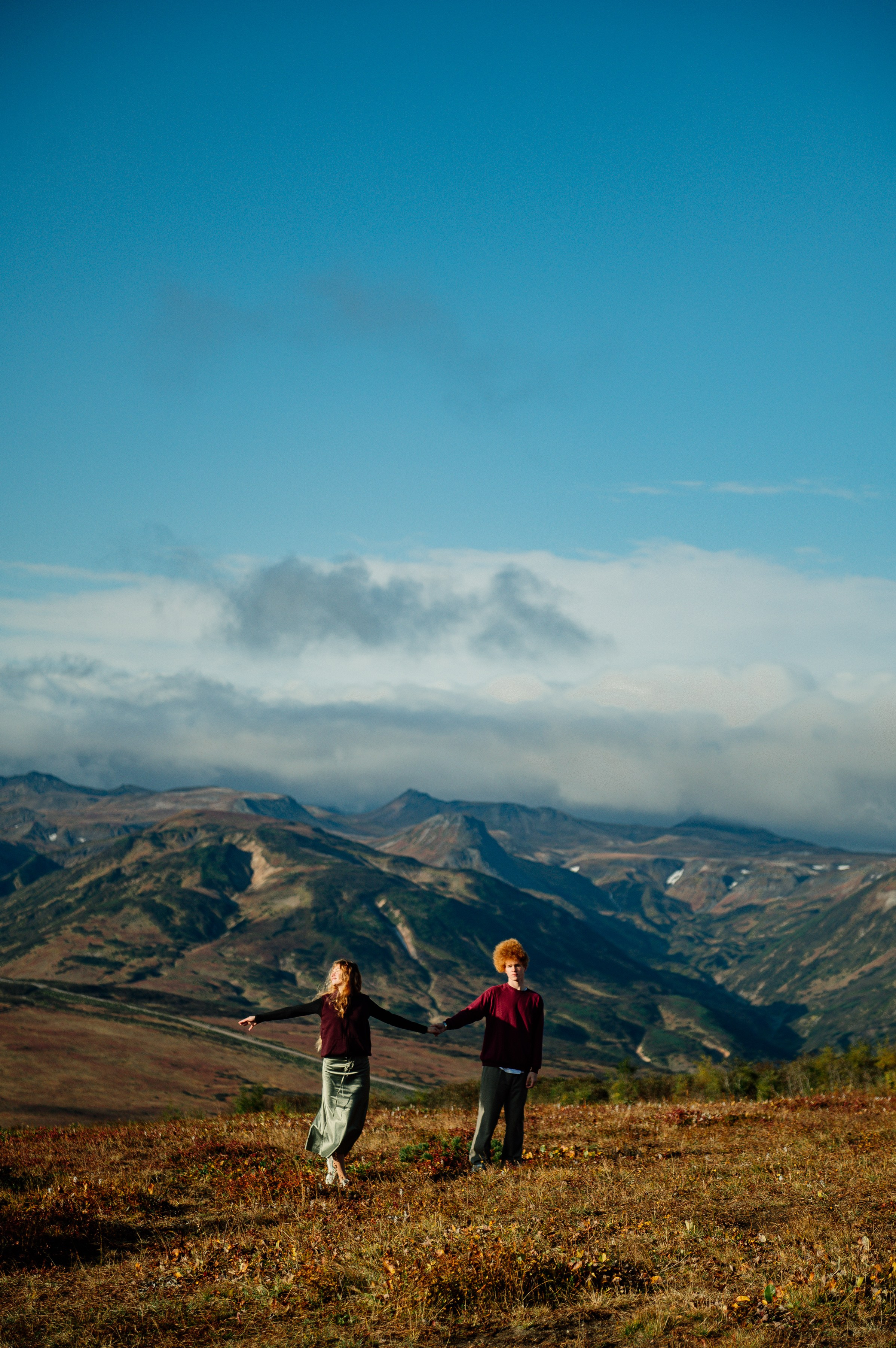 Парень и девушка держатся за руки на вершине холма, вокруг — горы и облака. Атмосфера свободная, вдохновляющая.