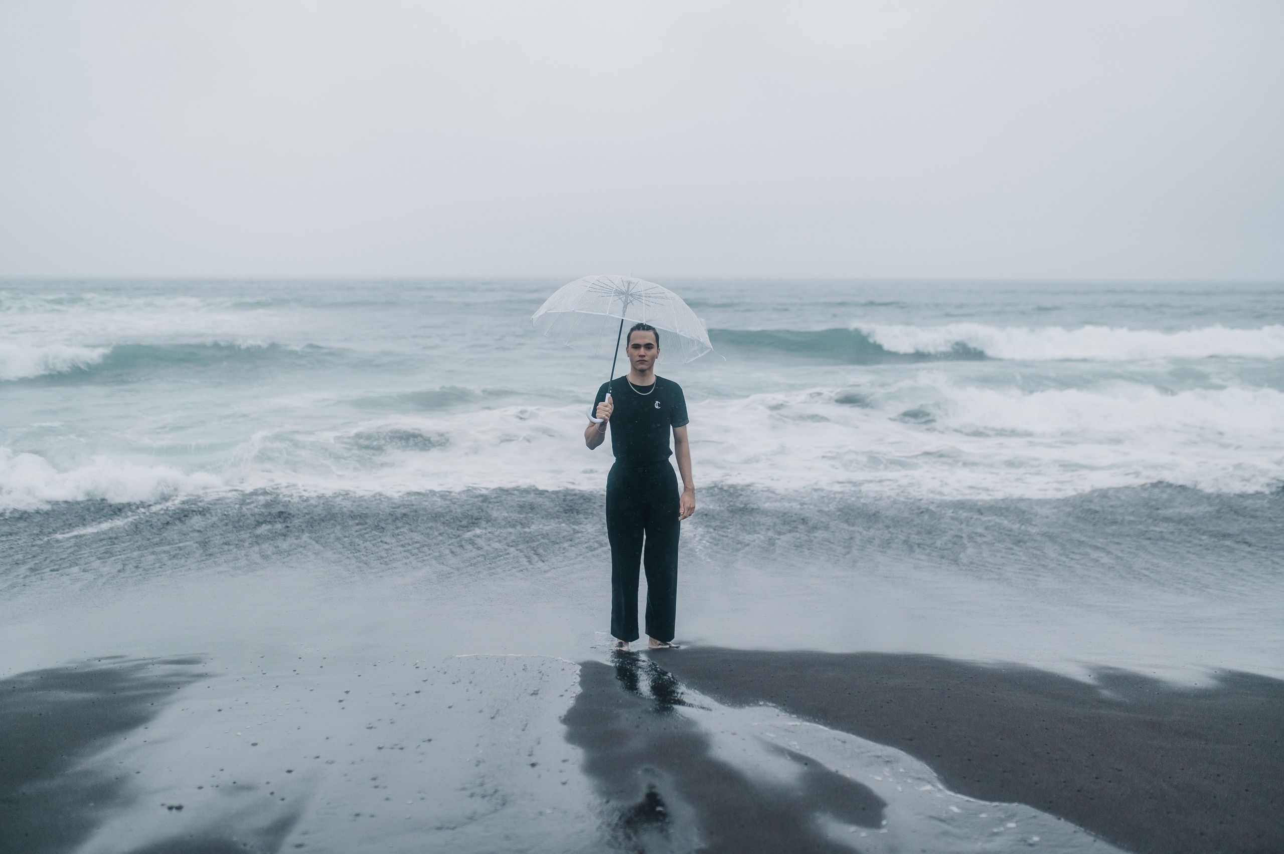 Парень в тёмной одежде стоит на мокром пляже с прозрачным зонтом, за ним бушует море. Атмосфера пасмурная, немного мрачная.