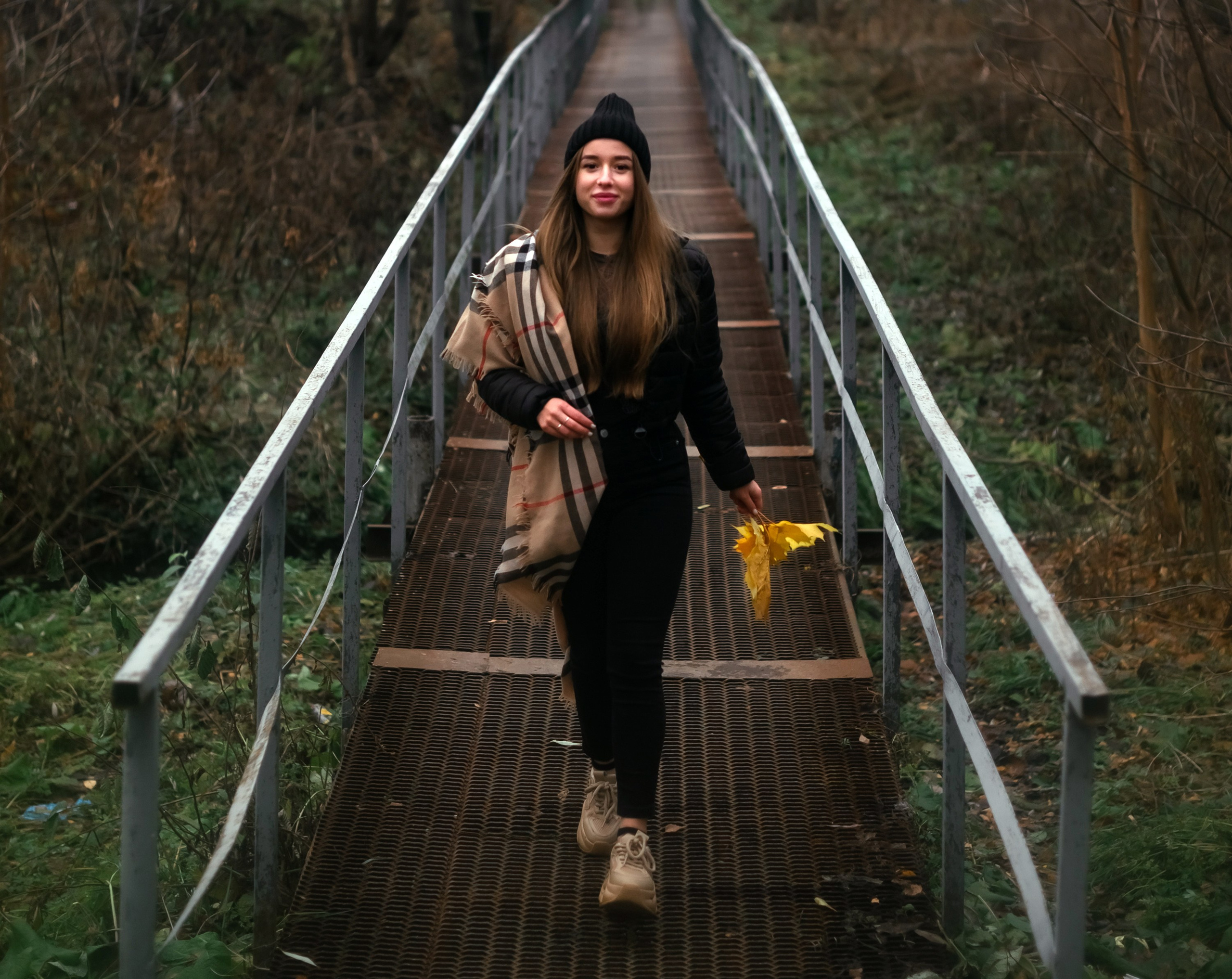 На фото портрет девушка осенними листьями идет по мосту.