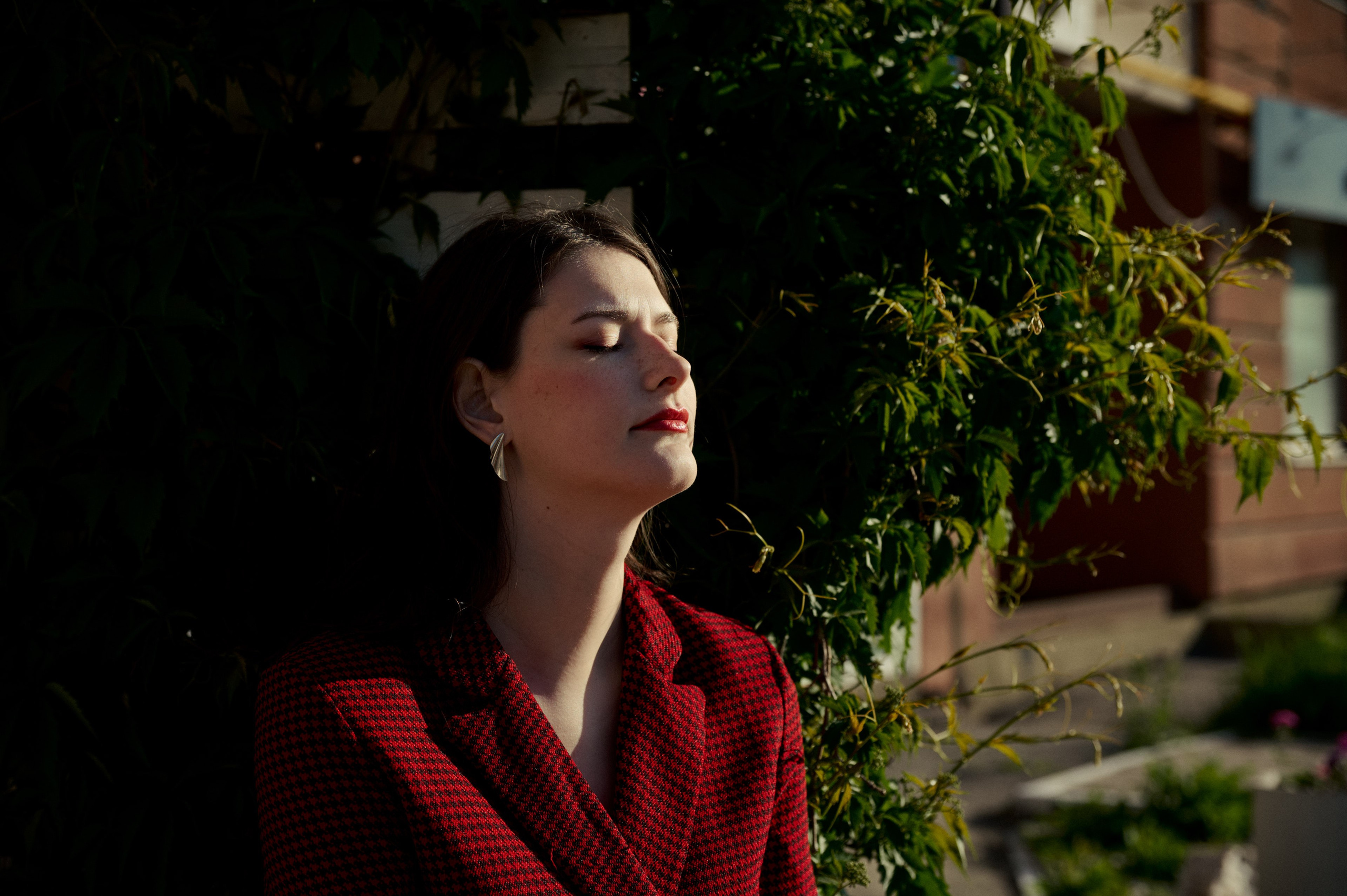 Уличная фотосессия в деловом стиле — женщина в бордовом закрывает глаза на фоне солнечной листвы.