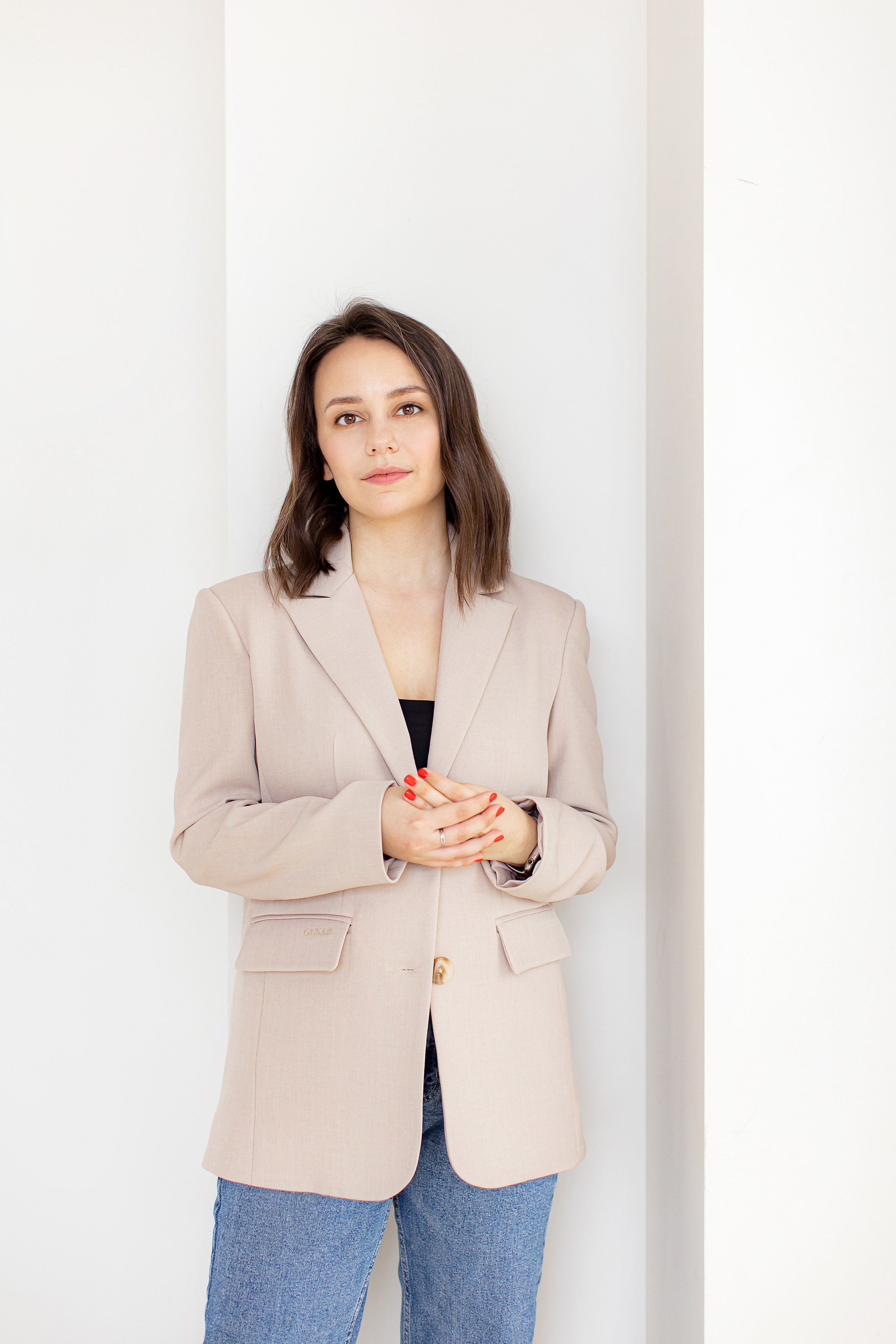 Девушка в бежевом пиджаке и синих джинсах стоит возле светлой стены