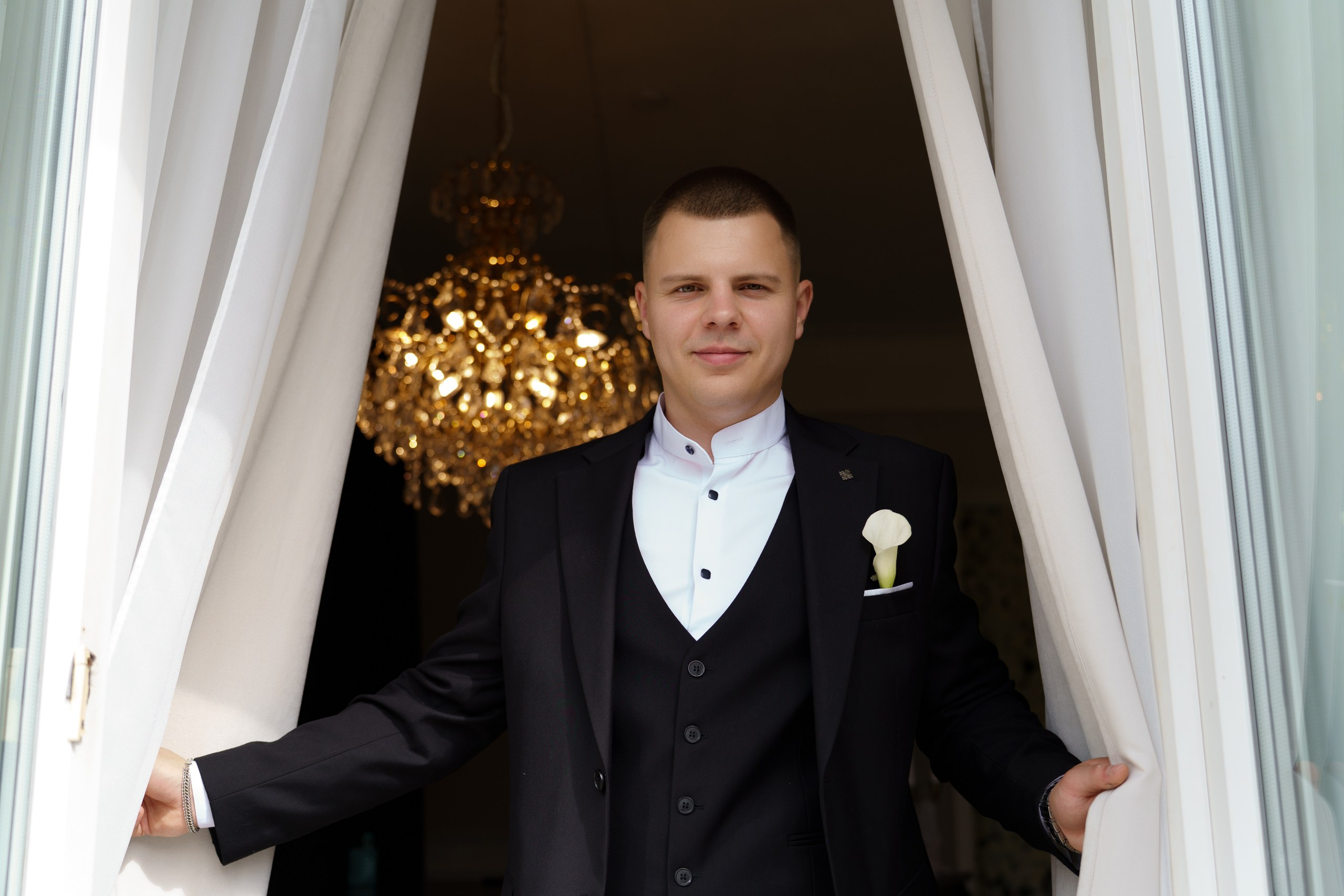 Свадебный портрет жениха: он одет в классический костюм с жилетом, белую рубашку и украшен бутоньеркой. Позирует, держась за шторы, на фоне роскошной люстры |свадебное фото на черном фоне Екатеринбург