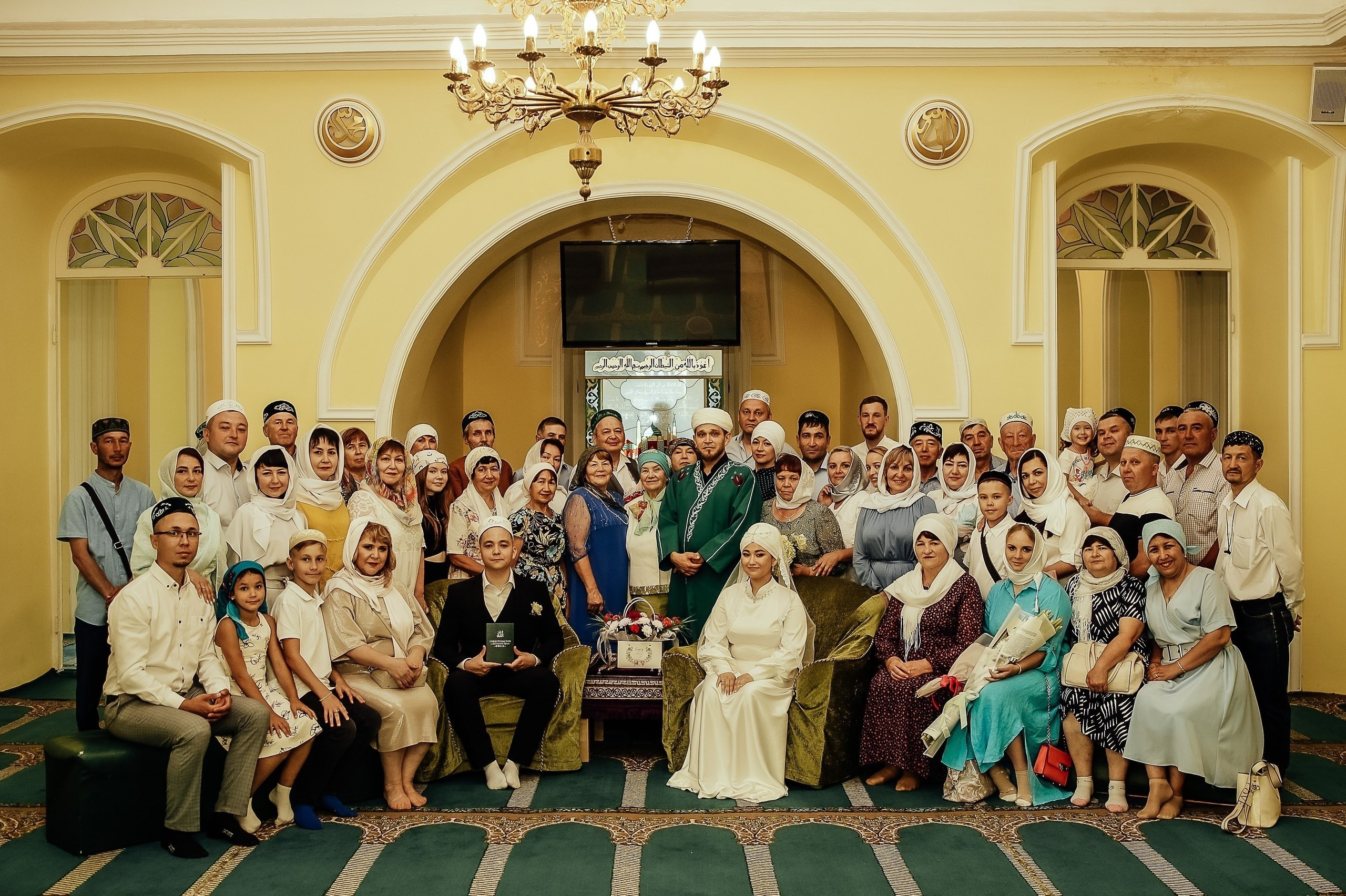 Большая семья и гости в традиционной одежде собрались в зале с люстрой. Атмосфера торжественная, радостная. Никах.