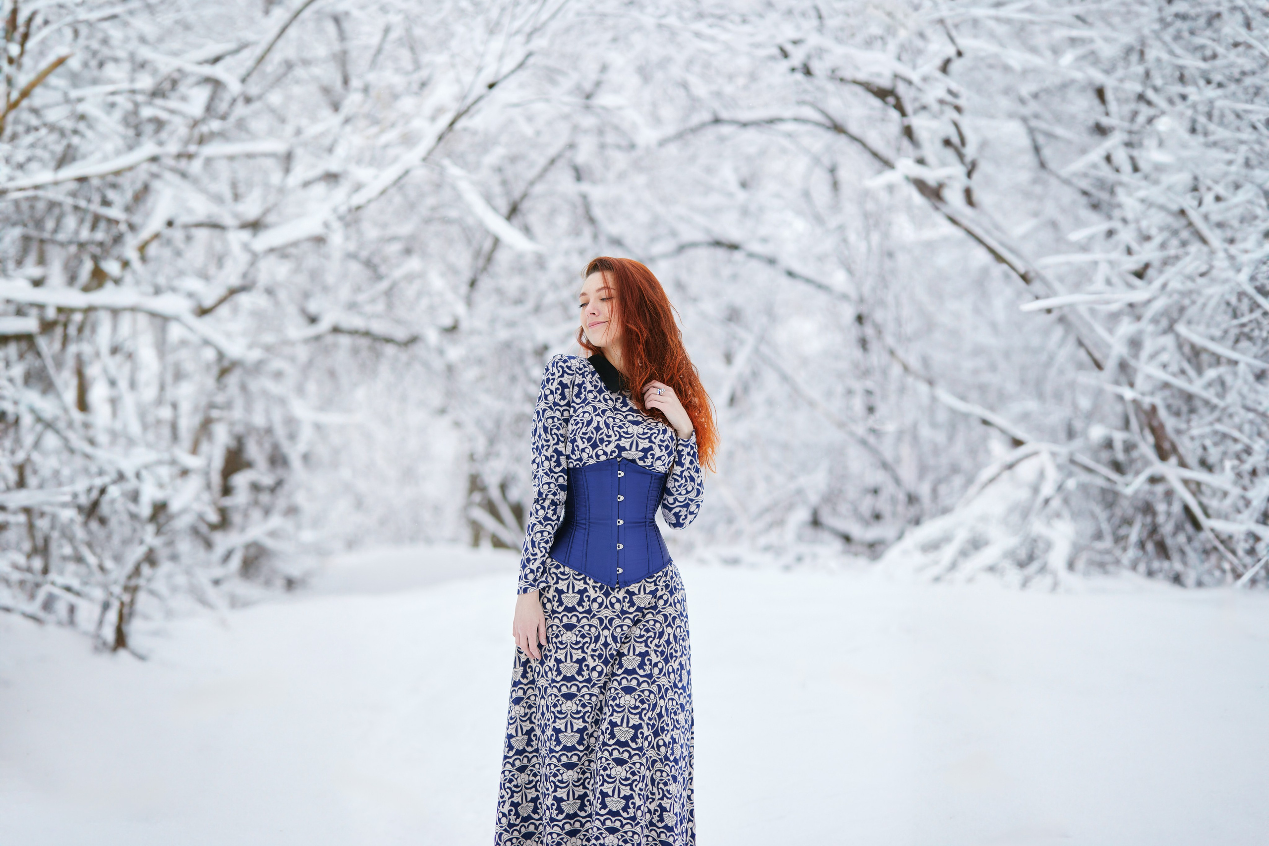 Красивая модель с длинными рыжими волосами улыбается и позирует на фоне зимнего леса