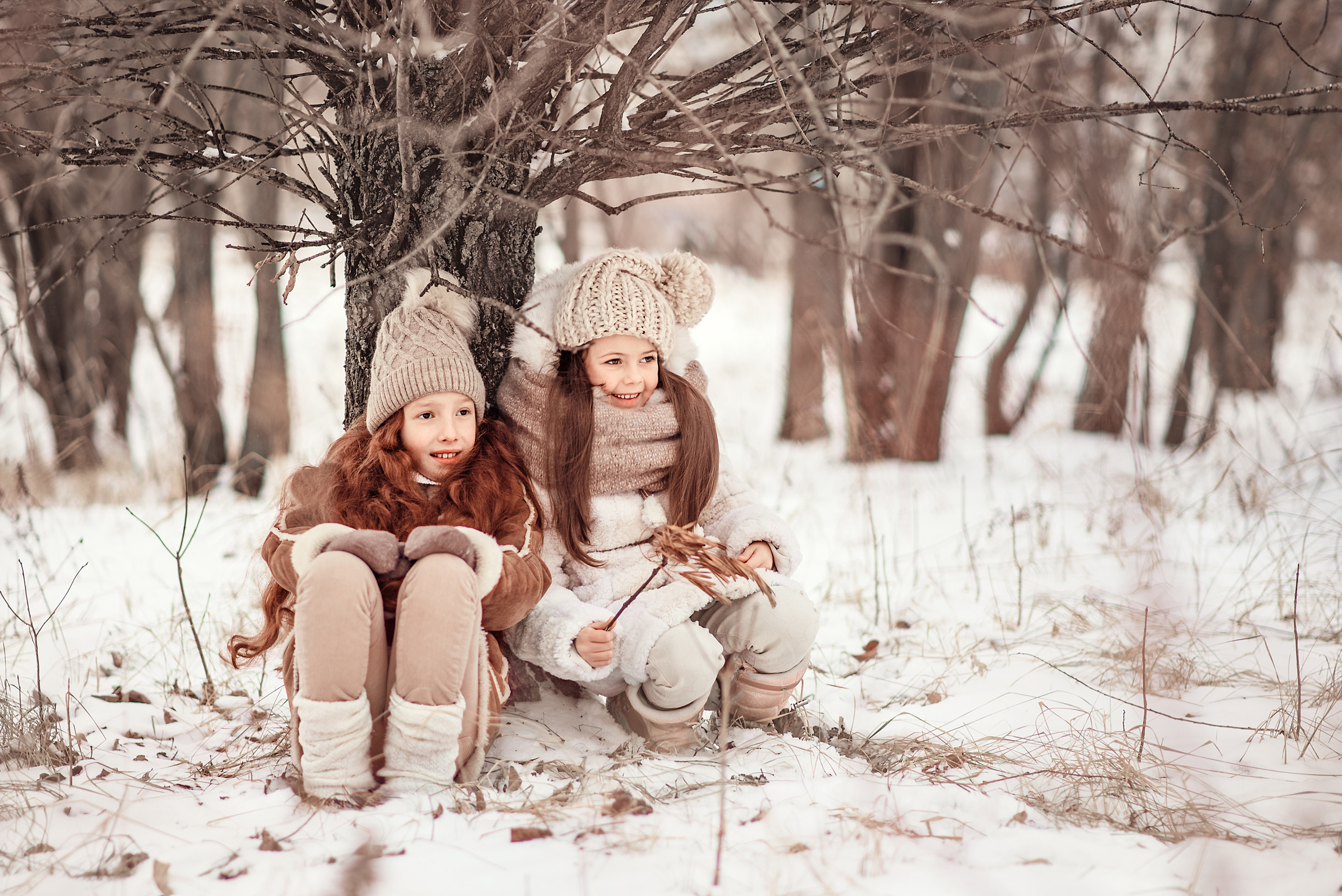 Две девочки сидят на снегу под деревом в зимнем лесу.