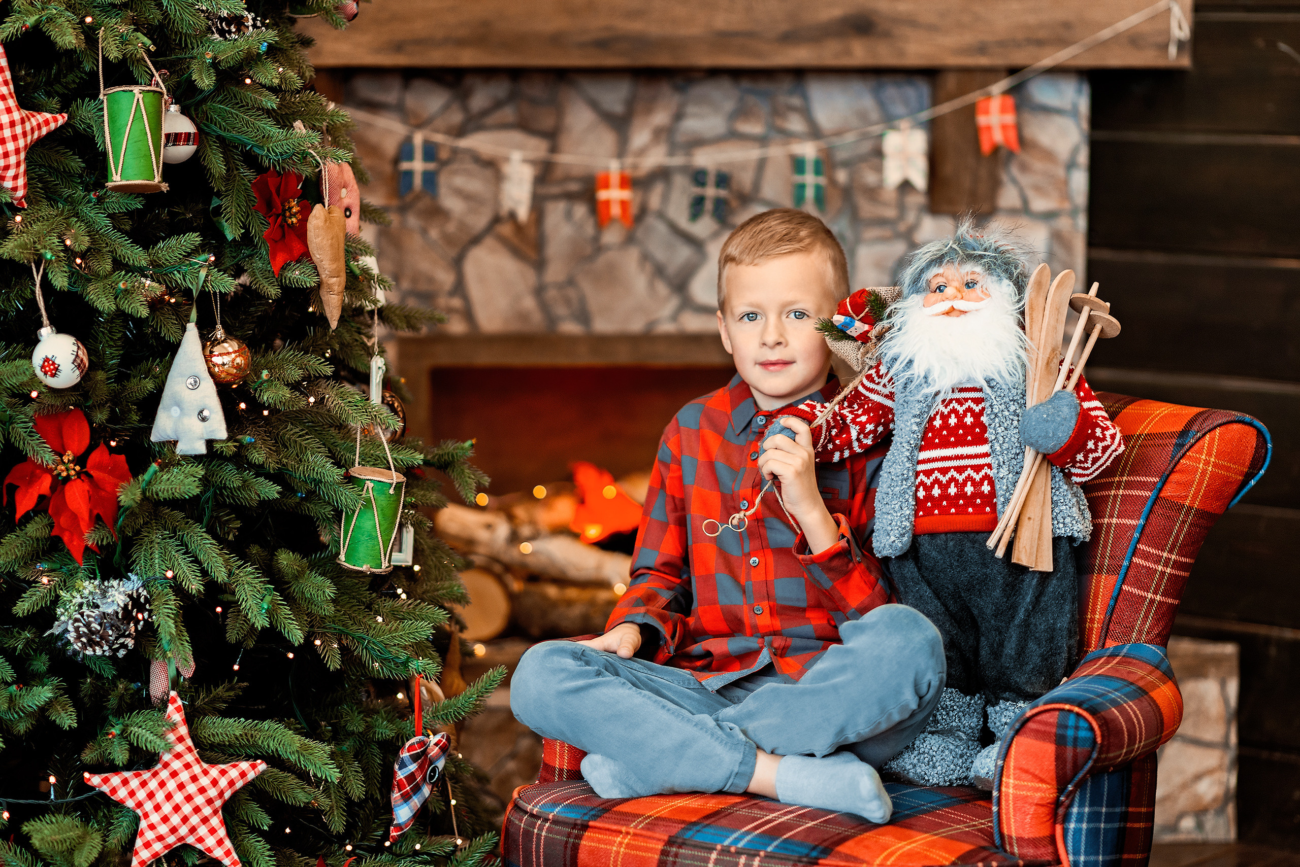 Мальчик сидит в кресле возле новогодней елки с игрушечным Санта-Клаусом.