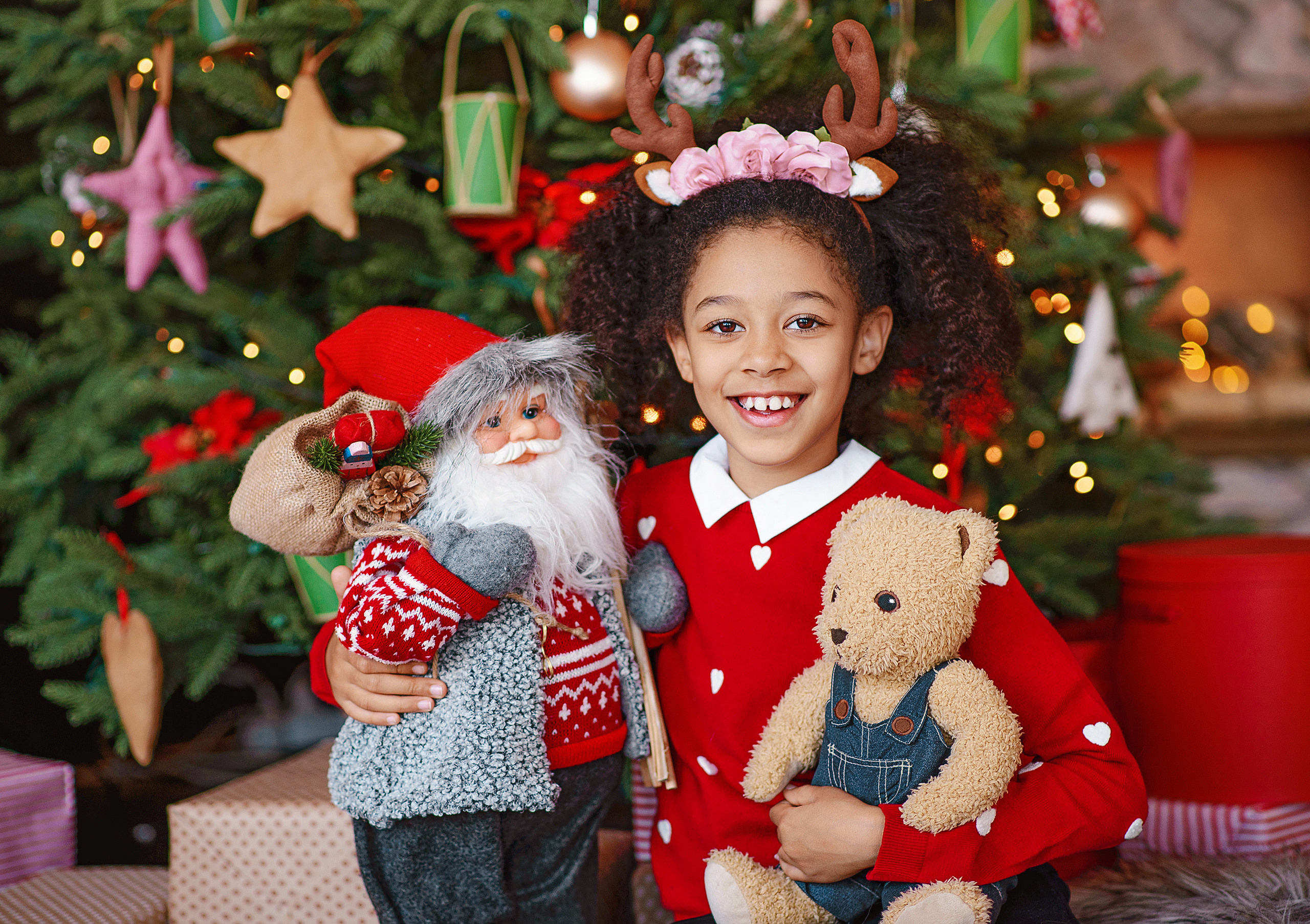 Девочка-мулатка в образе оленёнка позирует возле ёлки с игрушечным Санта-Клаусом и плюшевым мишкой.