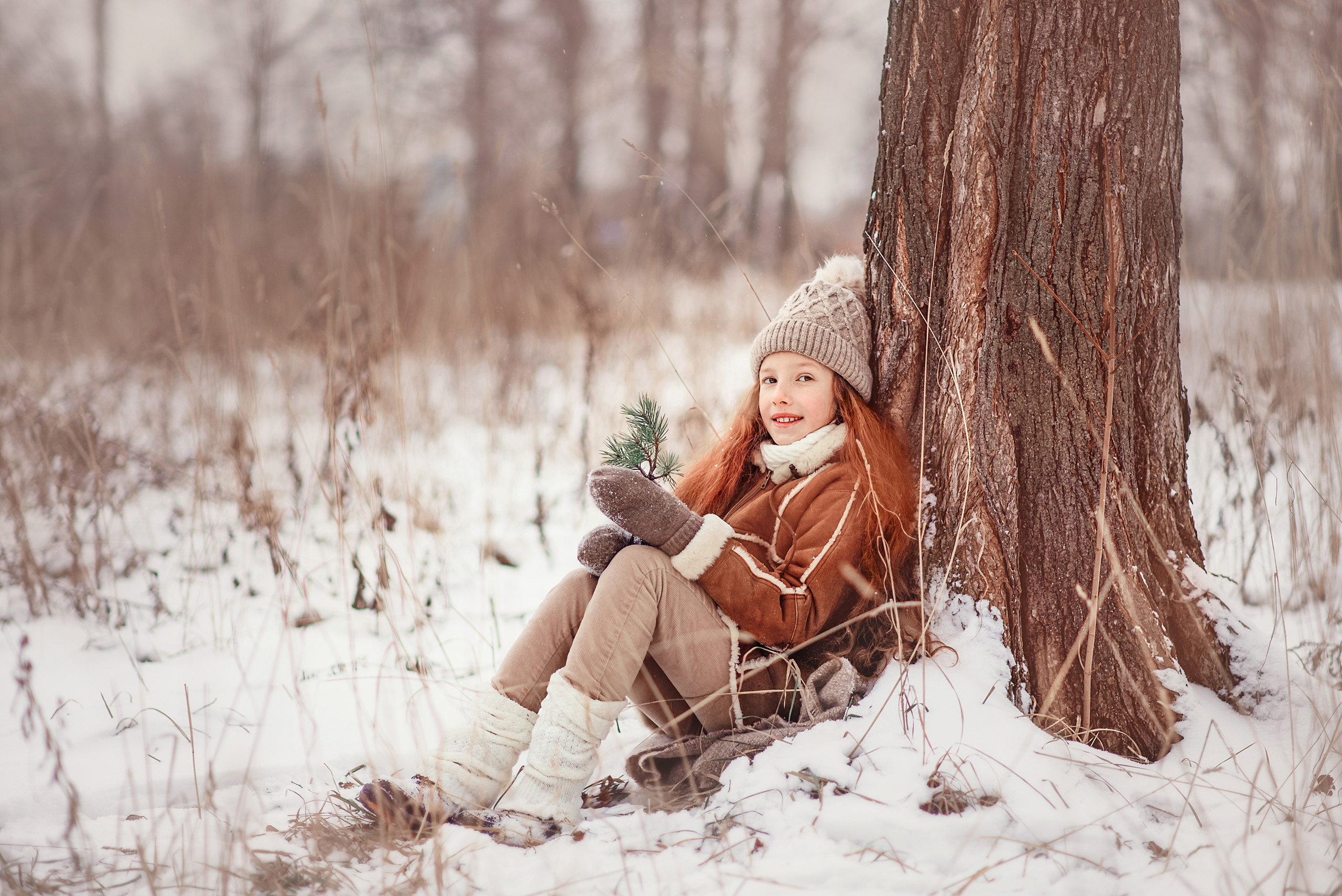 Девочка сидит под деревом на снегу и держит еловую ветку.