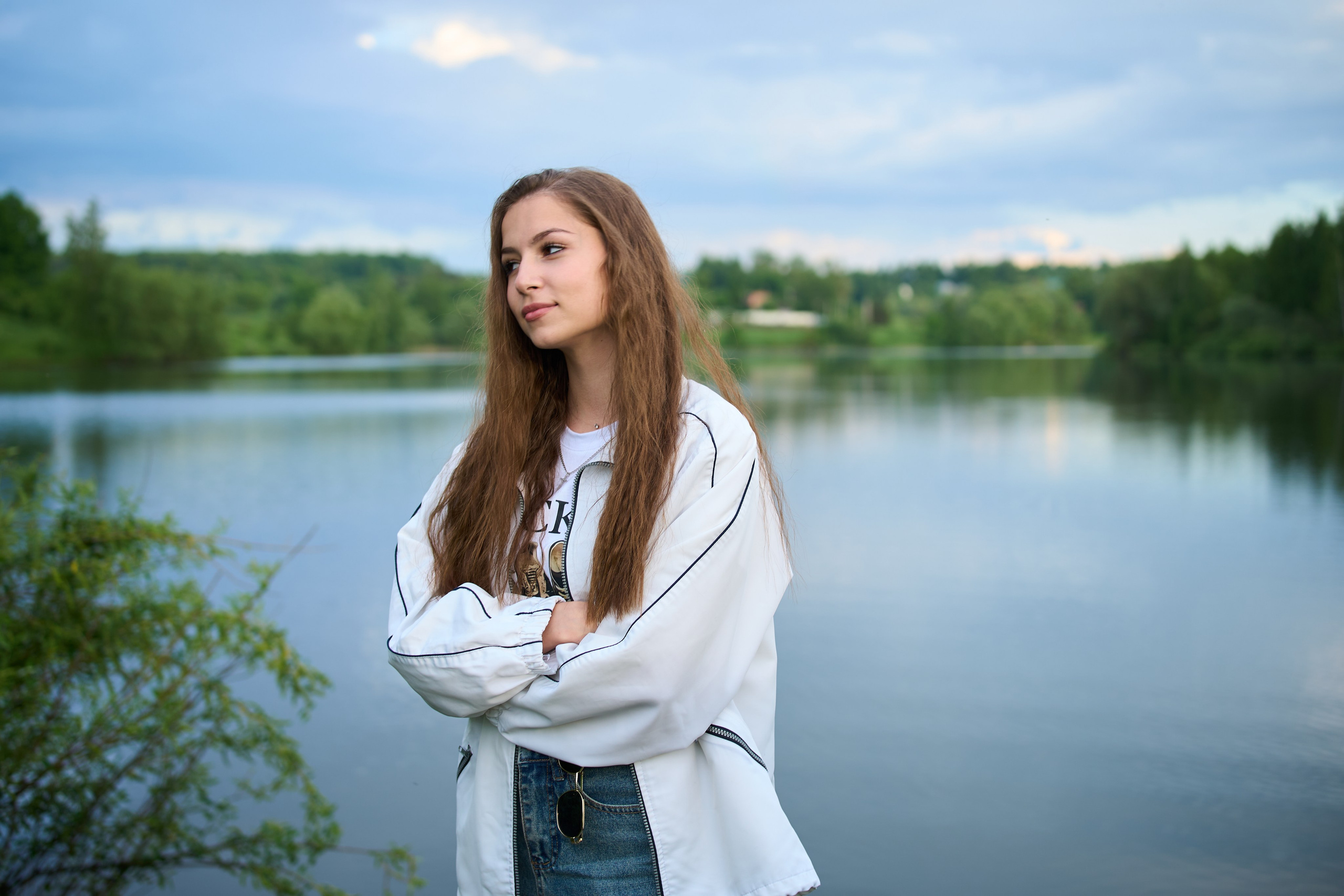 Женский портрет на природе — девушка с длинными волосами в белой куртке стоит у озера на фоне облачного неба